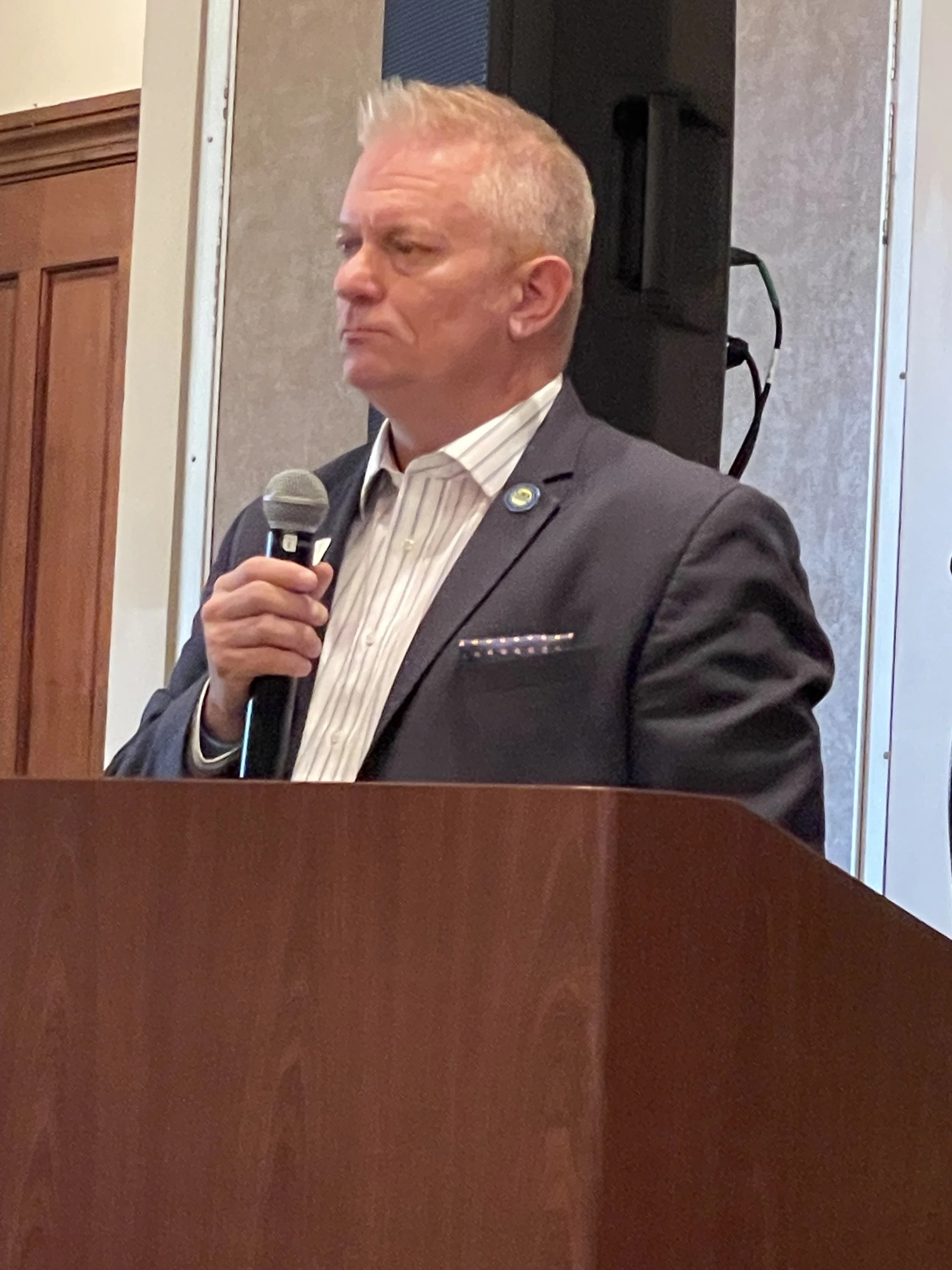 A man with short gray hair in a suit holding a microphone at a formal event, standing behind a wooden podium.