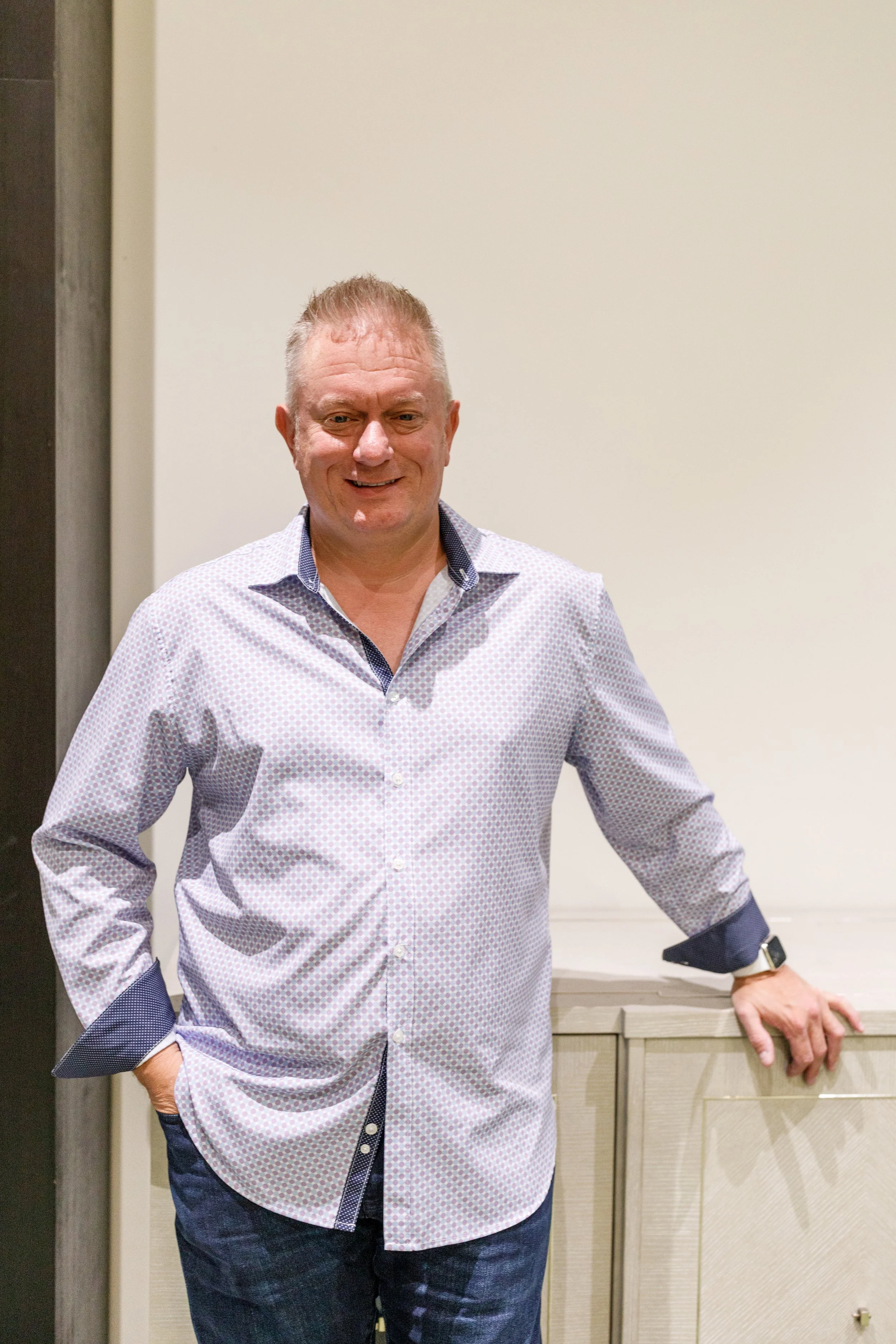 A smiling man with short hair wearing a patterned button-up shirt and dark jeans, standing indoors near a cabinet.