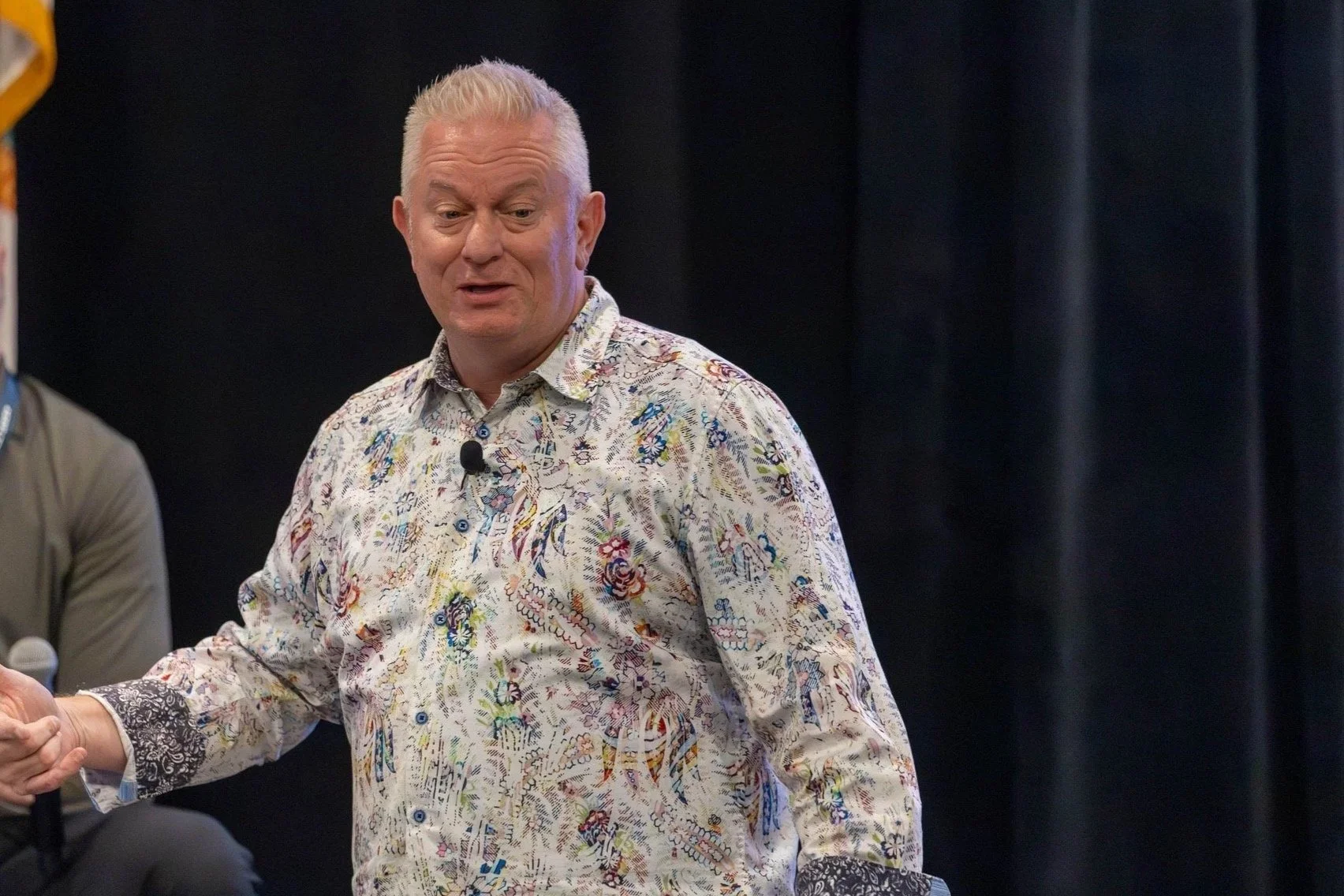 A middle-aged man with short gray hair wearing a colorful, patterned button-up shirt appears to be speaking or presenting, with a microphone clipped to his shirt, in front of a black curtain.