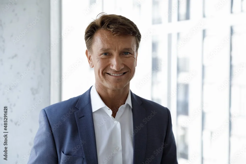 A man in a suit smiling, standing in front of large windows on a bright day.