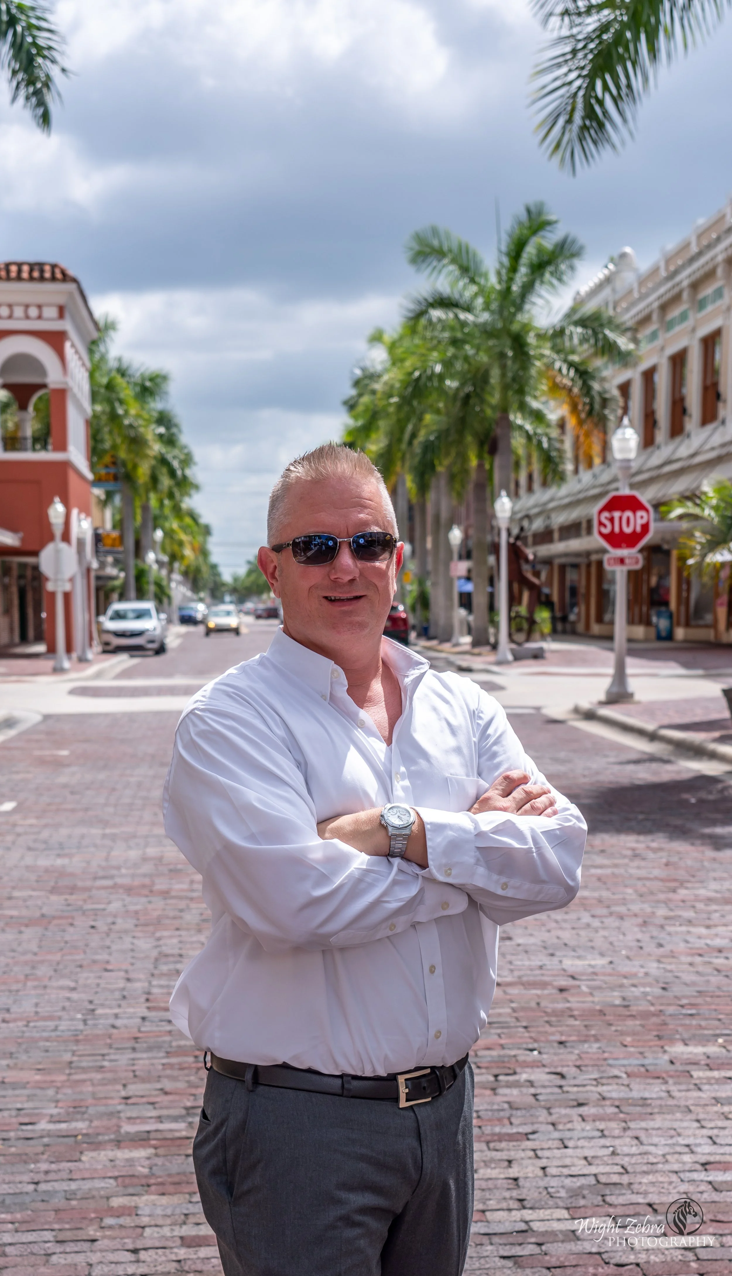 Jason Jakus National Speaker in a white dress shirt and dark pants with crossed arms, wearing sunglasses and a wristwatch, stands on a brick sidewalk in front of palm trees and buildings with a cloudy sky overhead.
