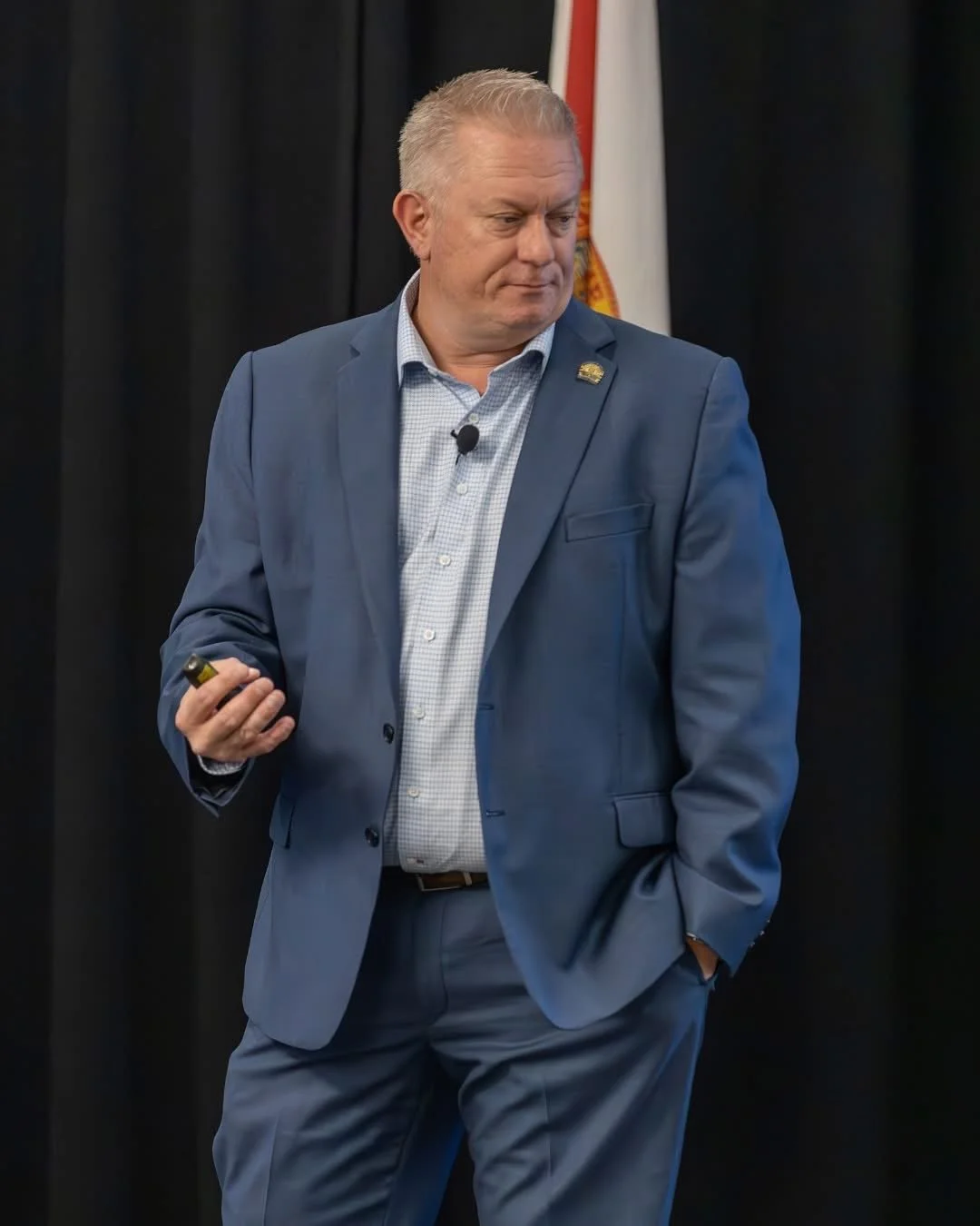 Jason Jakus National Speaker  in a blue suit giving a presentation, holding a remote, with a flag in the background.