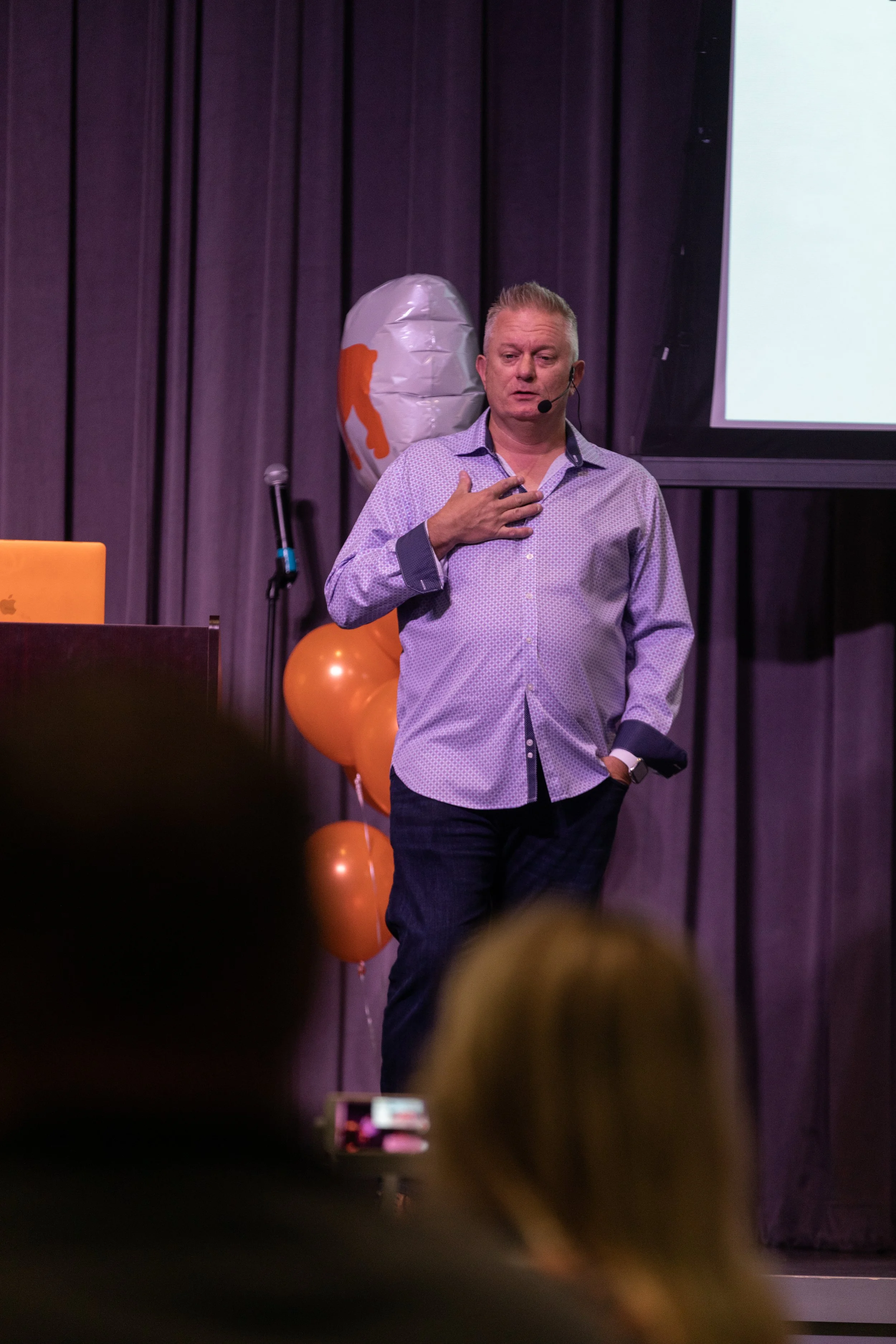 A man with short blond hair wearing a light purple button-up shirt standing on a stage with dark curtains behind him. He has his right hand over his chest and is speaking into a microphone headset. There are balloons and a large white screen in the background.