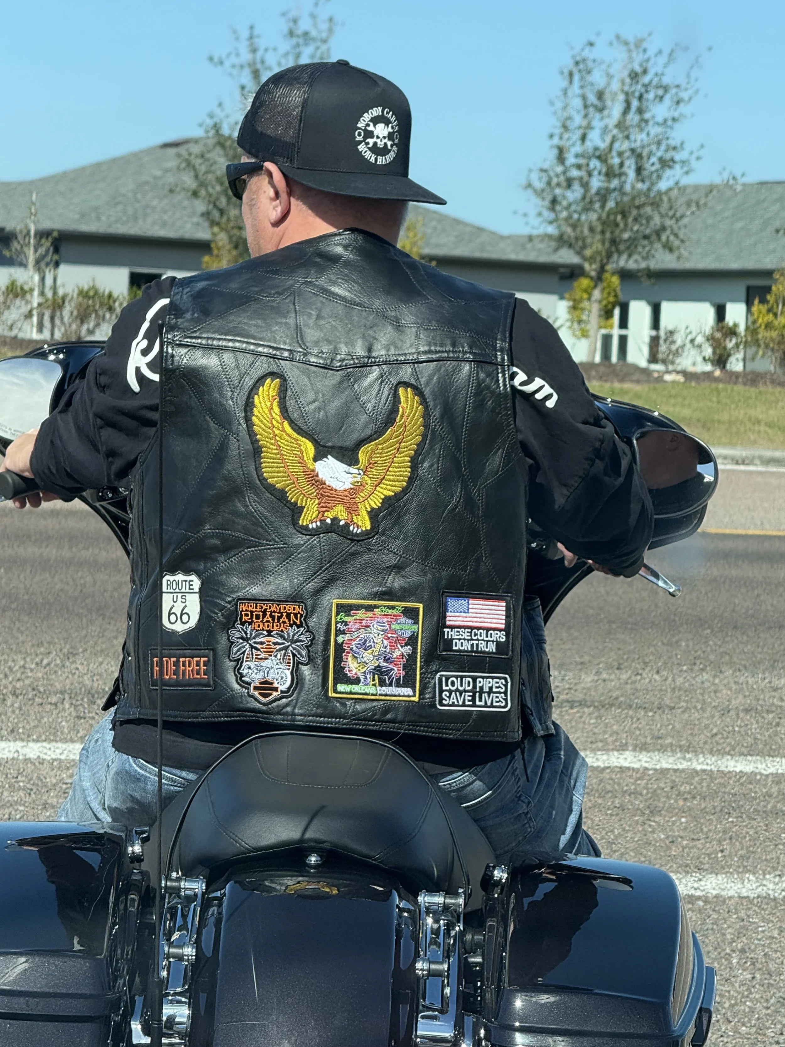 Jason Jakus wearing a black leather vest with patches, a black trucker hat with a skull logo, and sunglasses, riding a motorcycle on a street in front of residential houses with trees.
