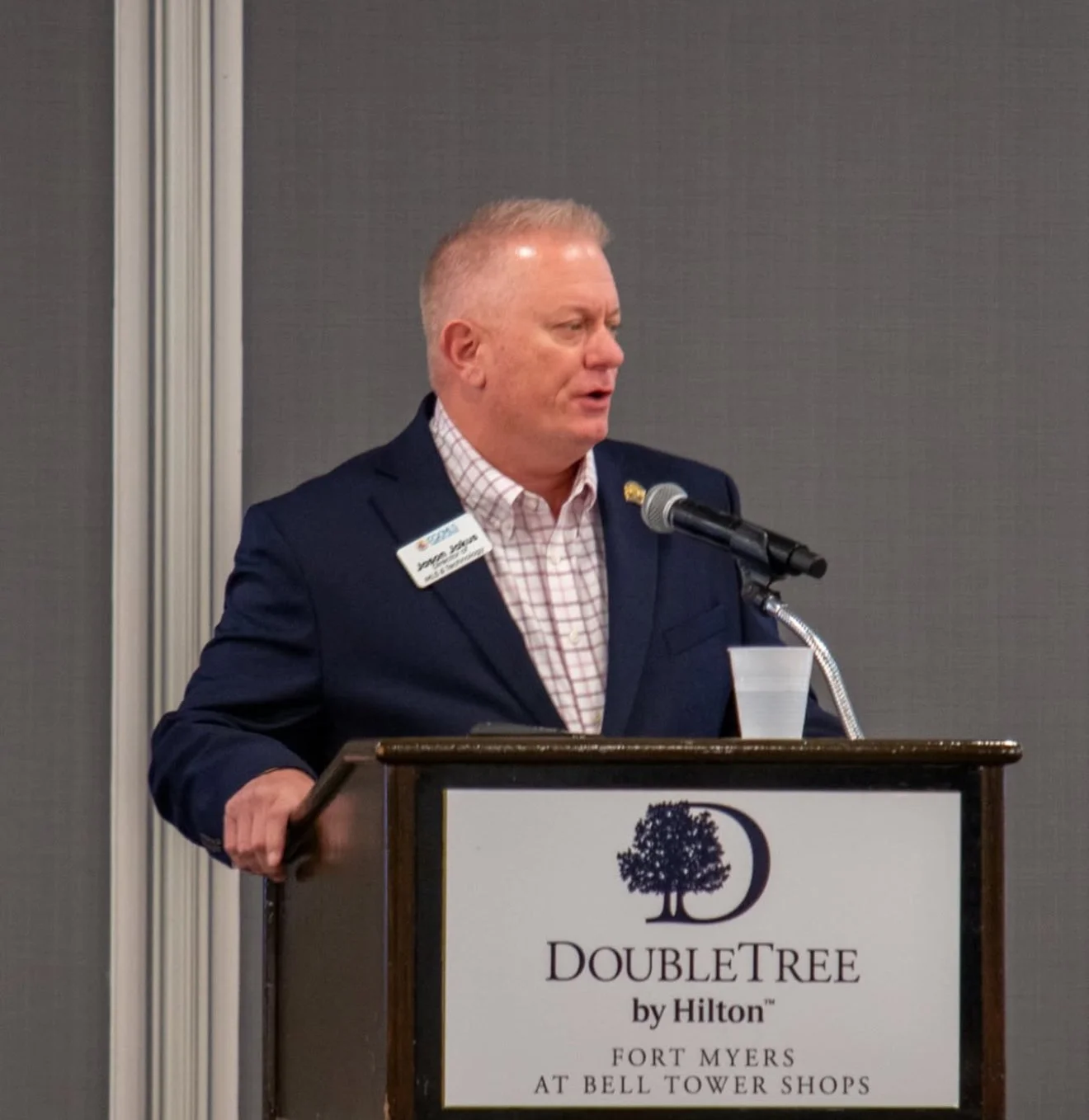 A man in a dark suit and checkered shirt speaking at a podium at a DoubleTree hotel event in Fort Myers, Florida.