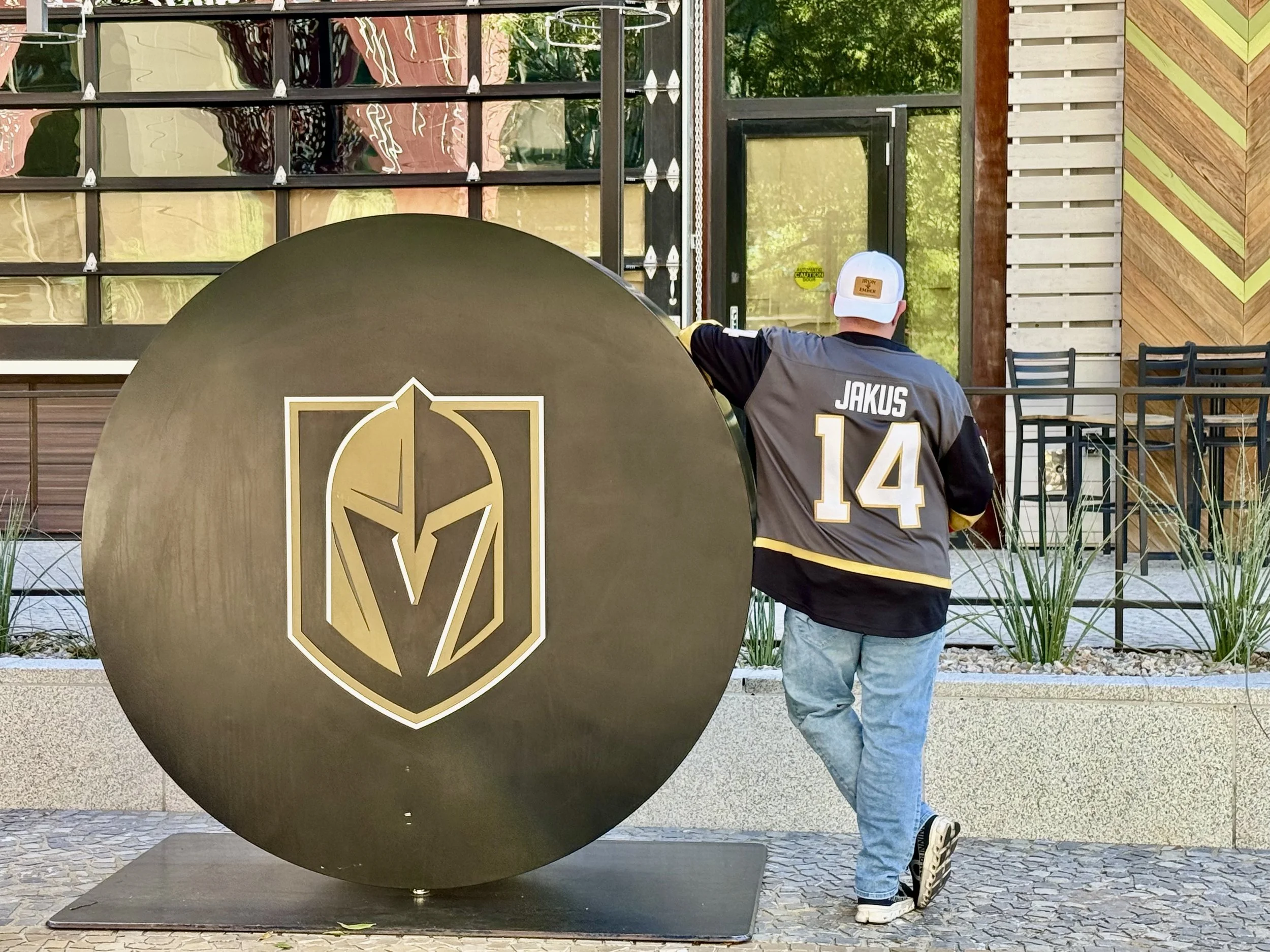 Jason Jakus wearing a gray sports jersey of the Las Vegas Golden Knights with the name 'JAKUS' and the number 14 on the back, standing next to the LVGK hockey puck at the T-Mobile Arena in Las Vegas.