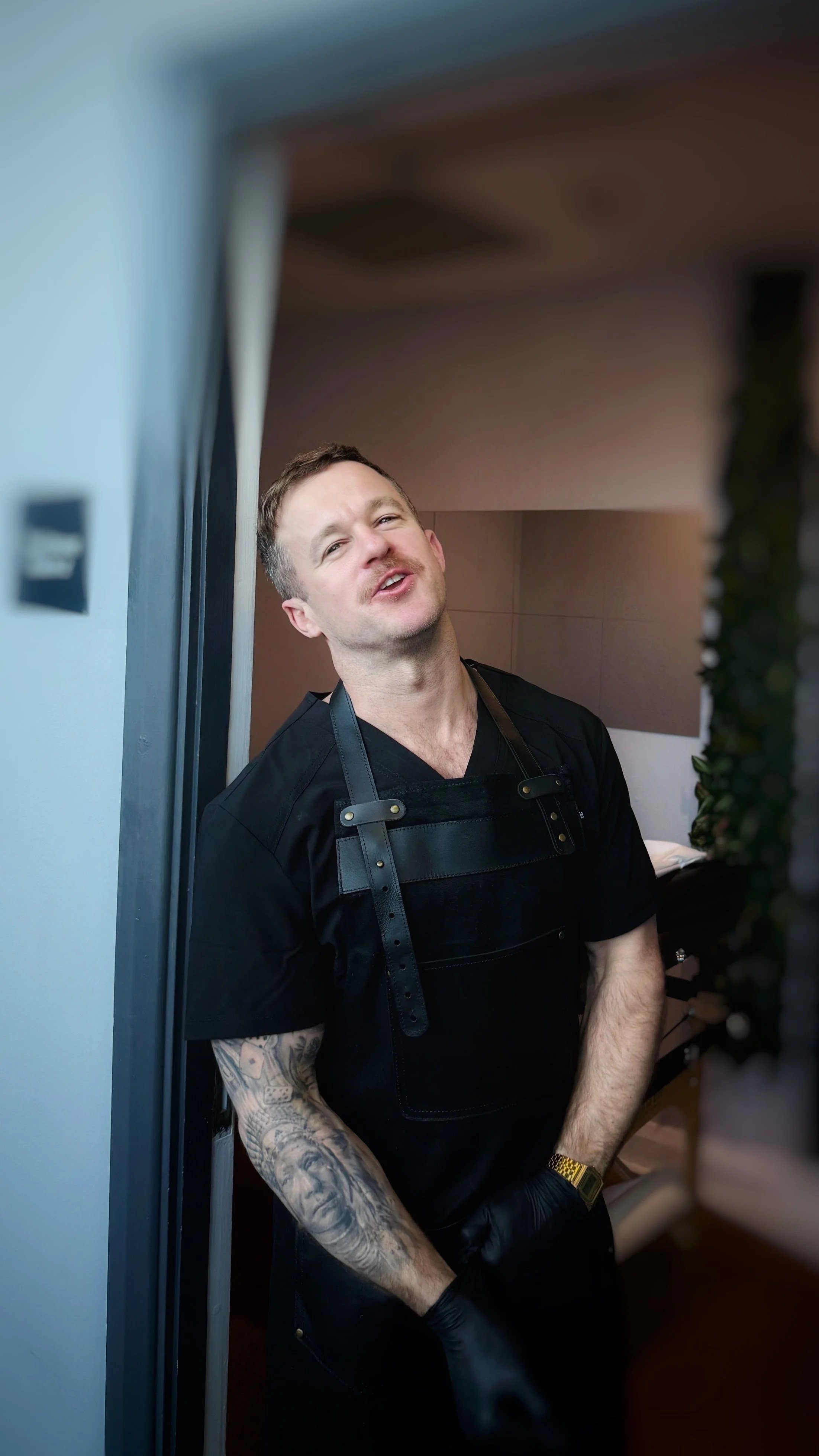 A man with a tattooed arm wearing a black uniform and a leather harness standing in a room with a plain brown and gray wall background.