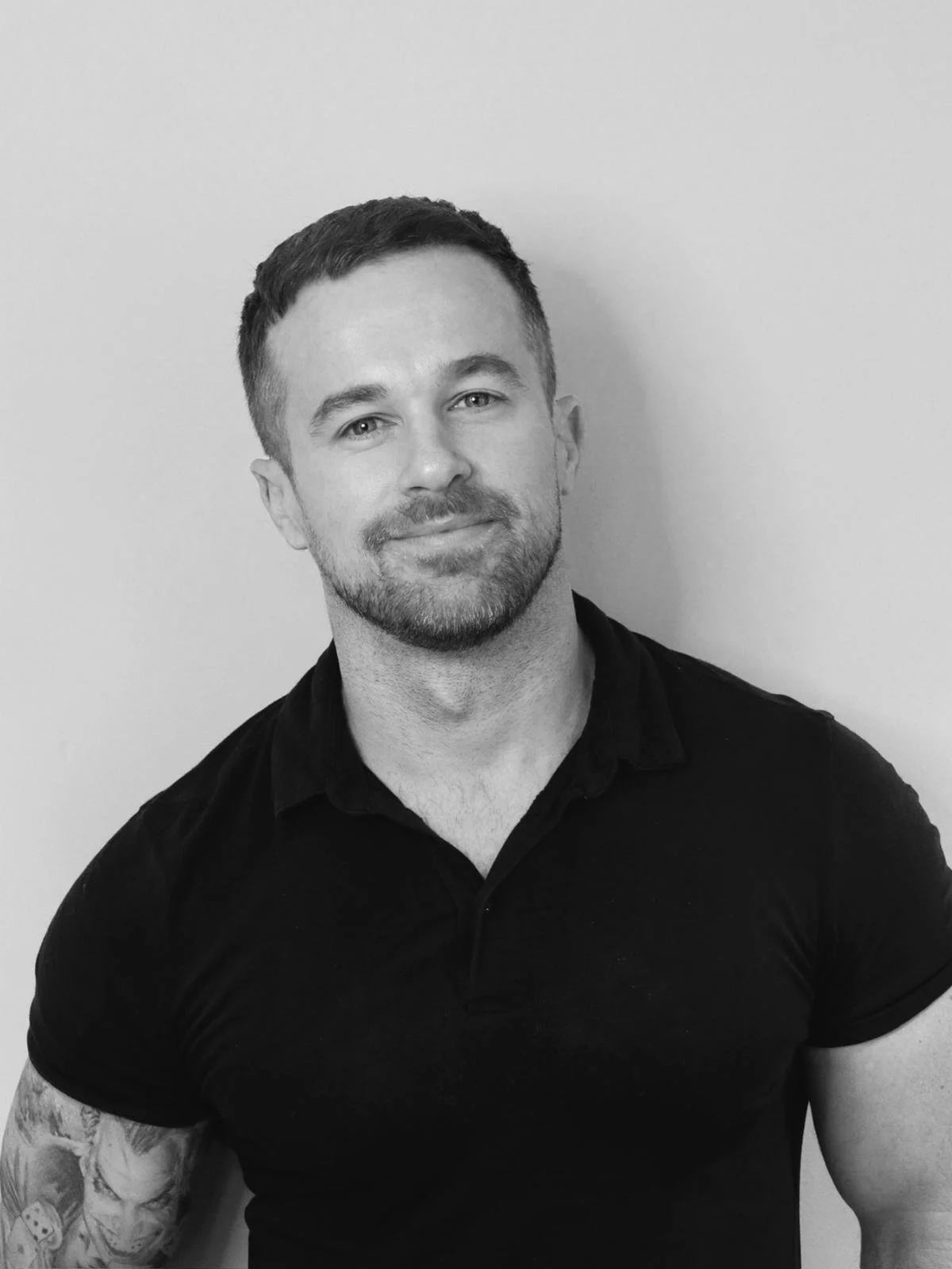 Black and white photo of a smiling man (Ed harbourne) with short hair and a beard, wearing a black shirt with rolled-up sleeves, standing against a plain wall.