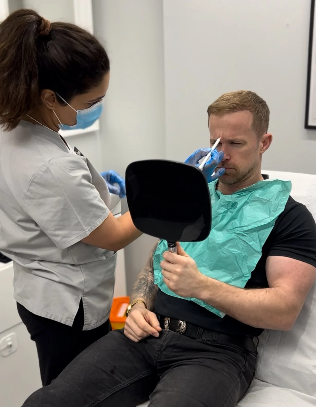A healthcare professional prepares to give a man (Ed Harbourne) skin boosters while he sits on a medical examination bed wearing a disposable green medical gown.