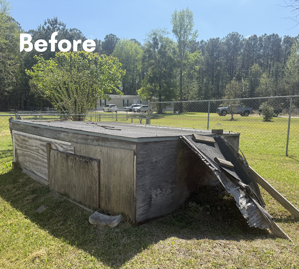 A damaged outdoor storage shed with a partially collapsed roof and burned exterior, situated on a grassy yard enclosed by a chain link fence.