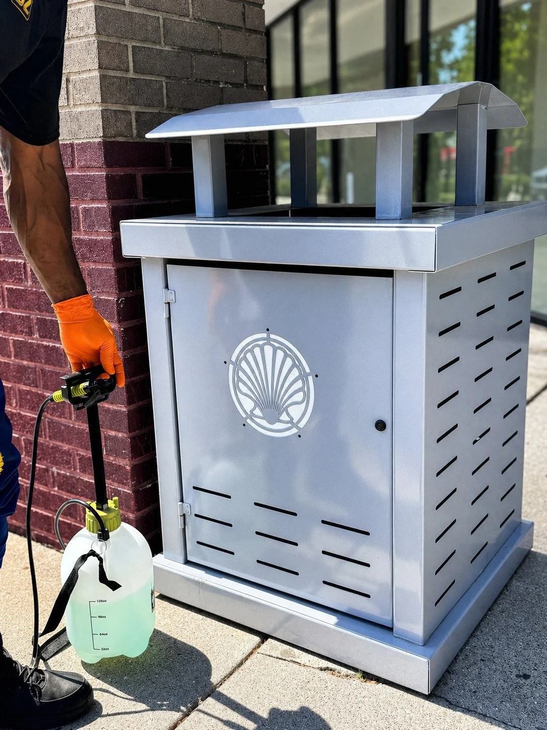 Person wearing black shirt and orange glove using a sprayer connected to a white tank, standing next to a silver outdoor trash or utility box with a shell logo, located outside a building with brick wall and glass window.