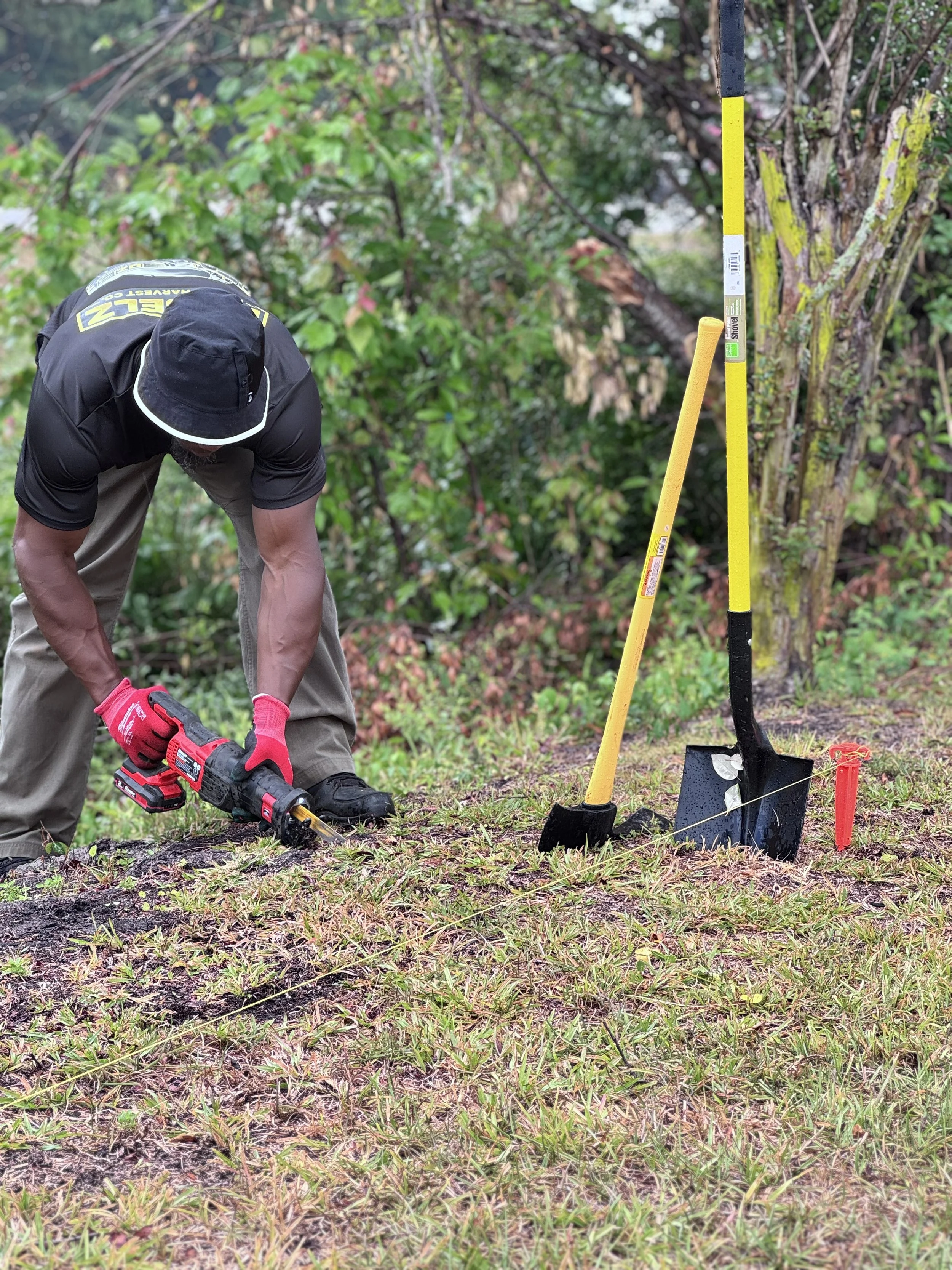 A person wearing a black hat, black shirt with yellow letters, khaki pants, and red gloves is working outdoors with a drill on the ground. There are yellow and black tools and markers, and soil being dug in a grassy area near a tree with green leaves