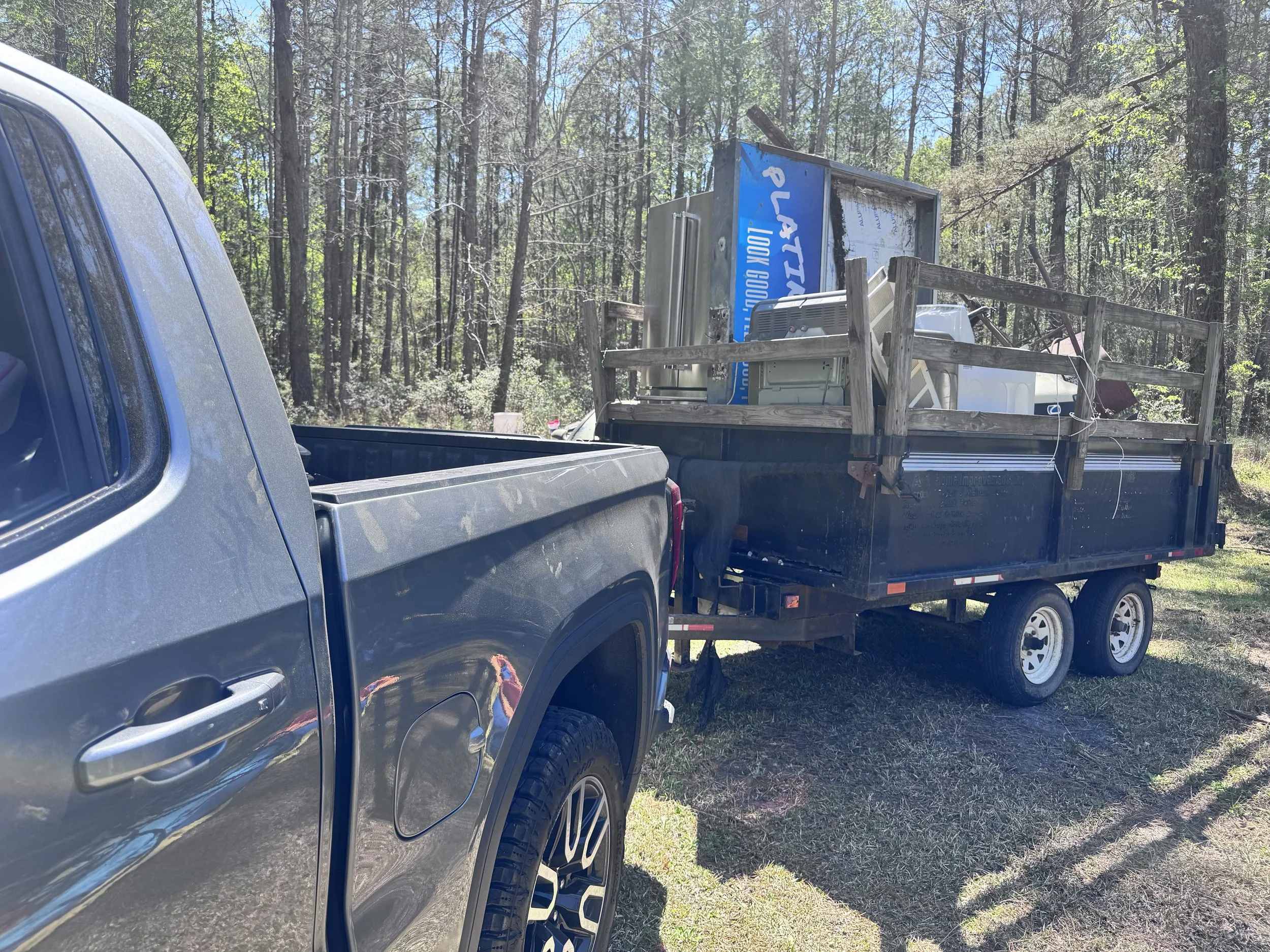 A pickup truck with a trailer attached, the trailer carrying a large ice vending machine, parked in a wooded area with tall pine trees and sunny weather.