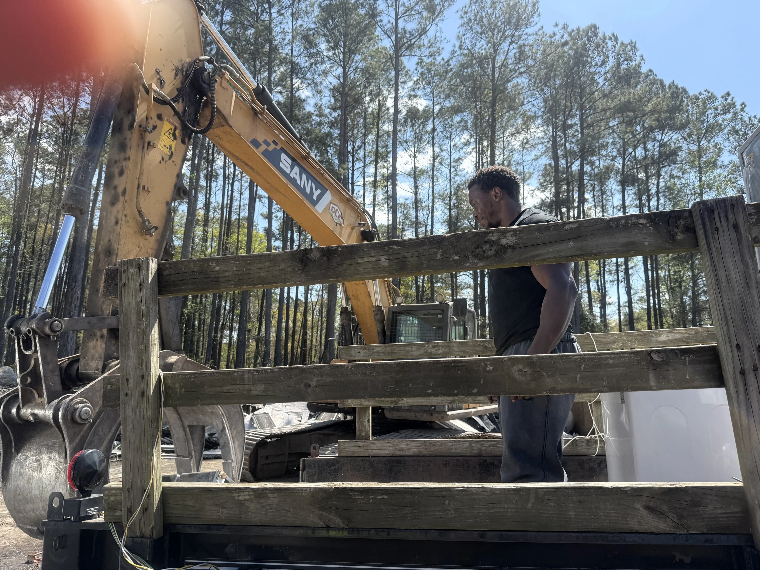 A man standing on a wooden platform, working with construction equipment in a forested area. A large yellow SANY excavator arm is visible above him, and the scene is outdoors on a clear day.