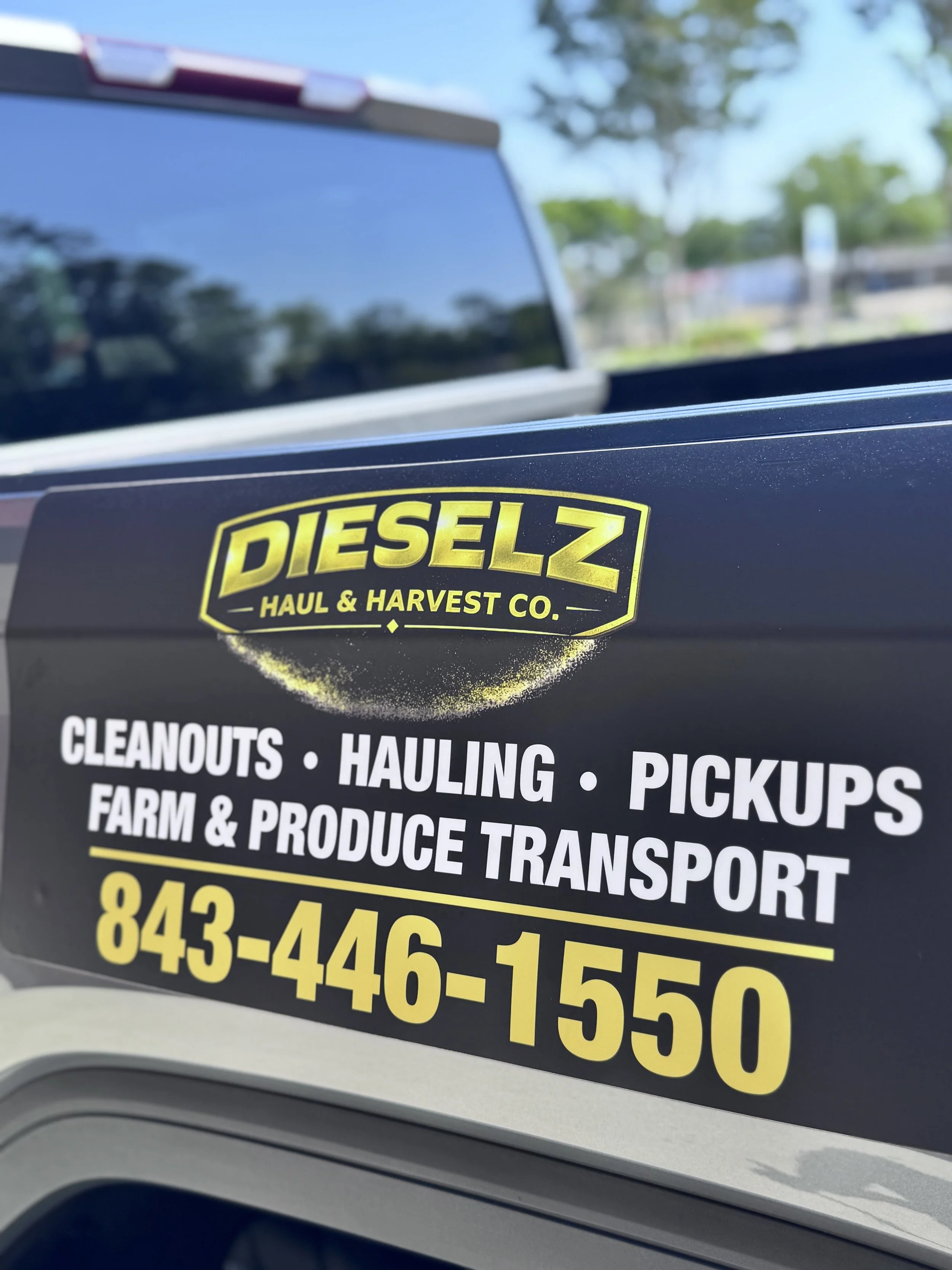 Close-up view of a black sign with yellow and white text advertising services for a haul and transport company, including 'Cleanouts, Hauling, Pickups, Farm & Produce Transport,' and a phone number. The background shows part of a vehicle window and a blurry outdoor scene with trees and blue sky.