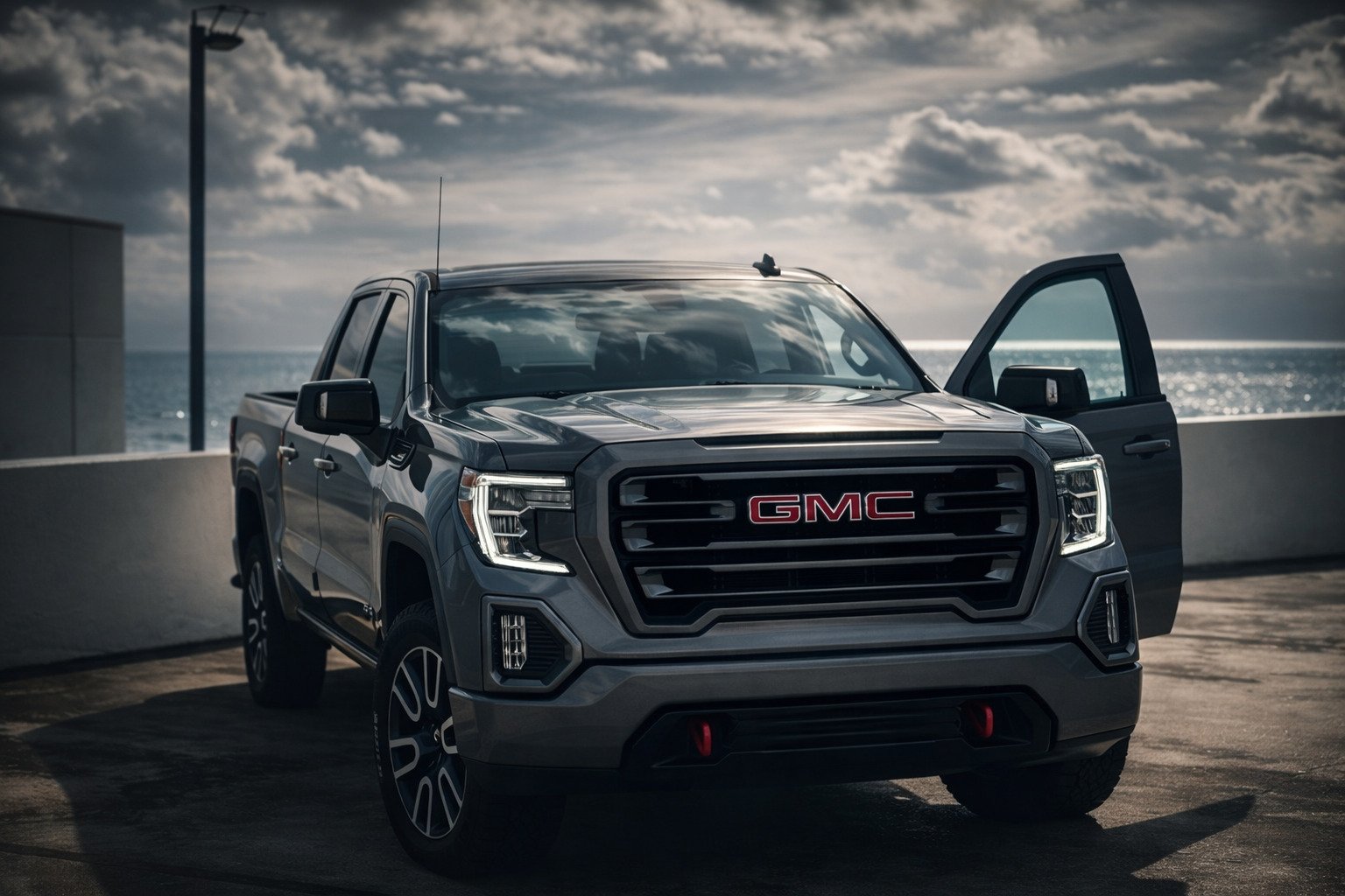 A dark gray GMC pickup truck parked on a rooftop parking area near the ocean, with the driver's side door open and a cloudy sky above.