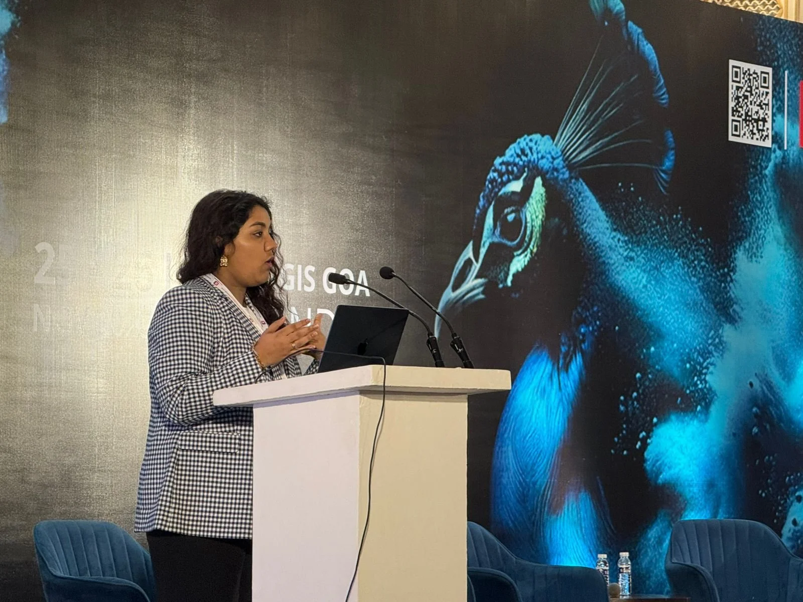 A woman in a checkered blazer speaking at a podium with a laptop, behind a large poster of a peacock.
