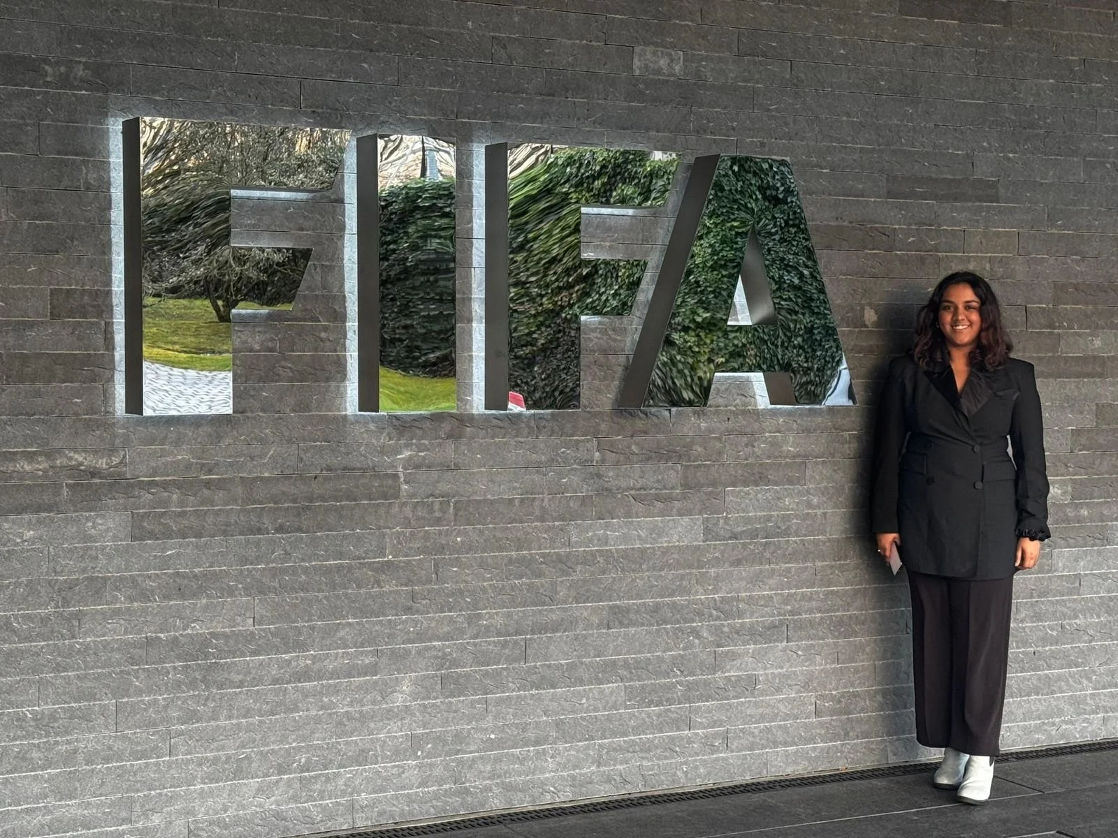 Woman in black blazer and black pants standing beside a large mirror with the word 'FIFA' on a gray brick wall.