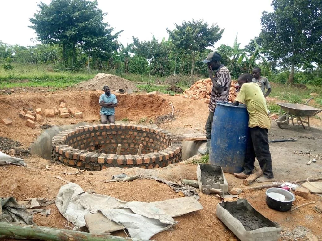 Group of five men working on construction of a circular brick structure outdoors. One man stands inside a partially excavated area, while the others stand around, some observing and others preparing tools. Construction materials and tools are scattered around.