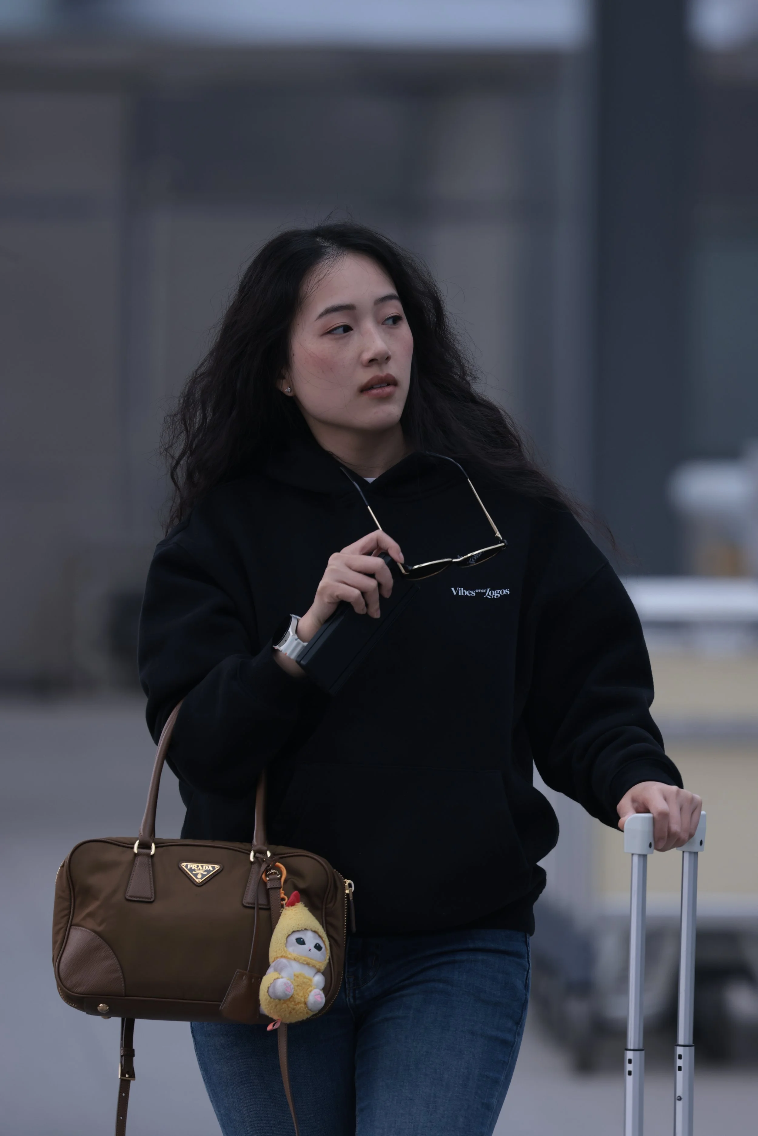 A woman with long dark curly hair wearing a black hoodie and blue jeans, carrying a brown Prada handbag with a plush duck toy attached, holding a rolling suitcase handle, walking outdoors with a gray, blurry background.