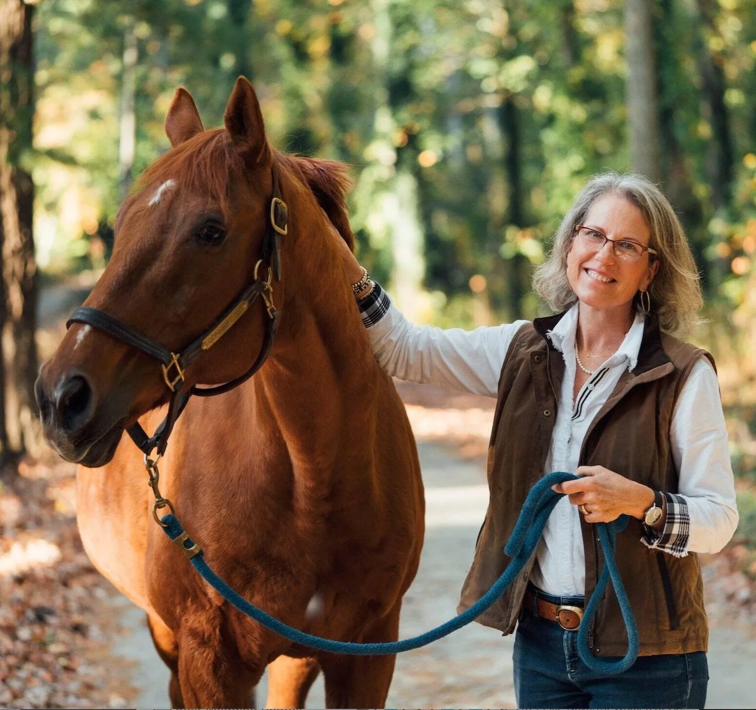 A woman with glasses, gray hair, and a smile, standing outdoors in a wooded area, holding a blue lead rope attached to a chestnut horse with a bridle, during autumn.