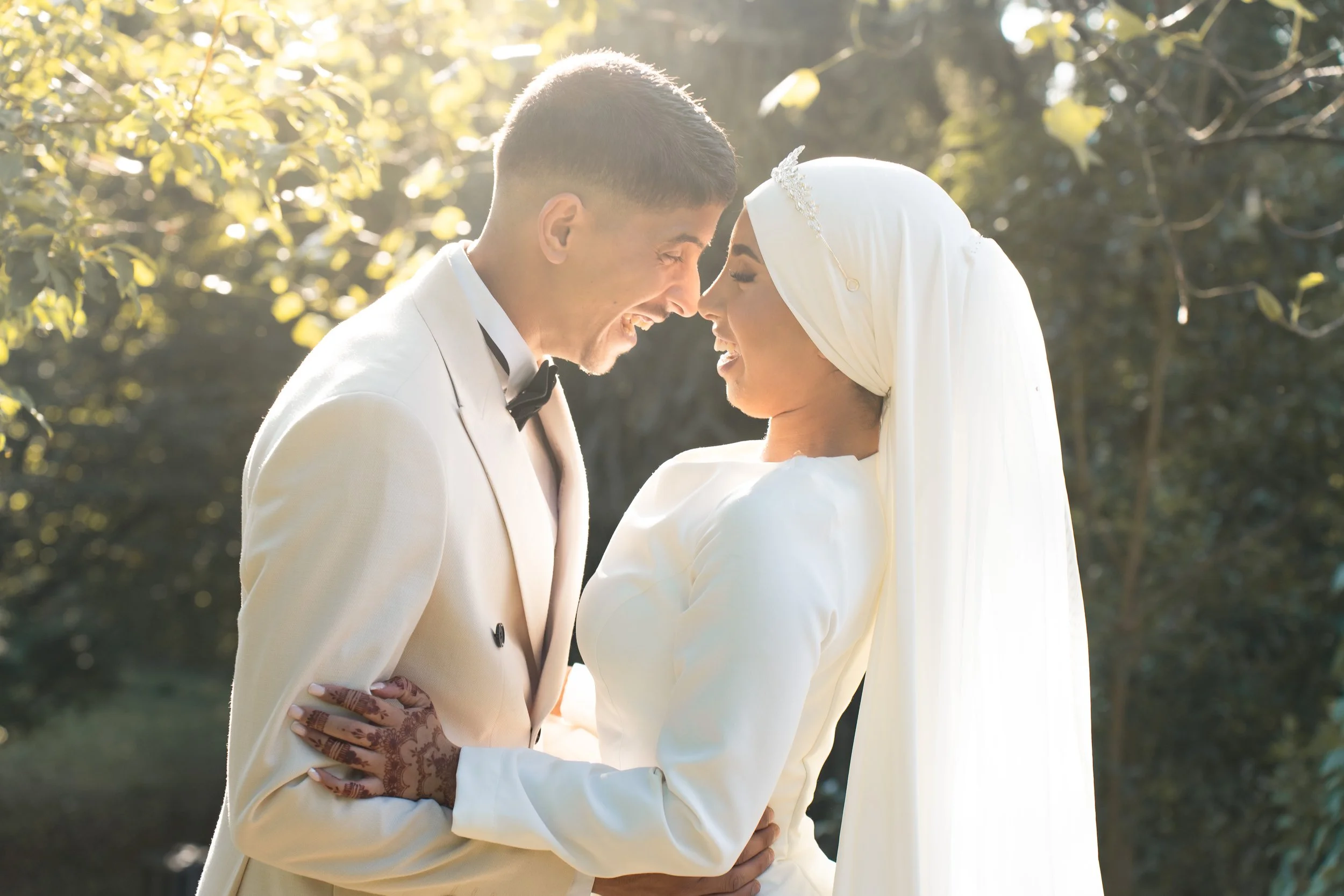 Un couple de mariés qui se tient face à face, souriants, lors d'une cérémonie de mariage en plein air, sous un arbre ensoleillé.
