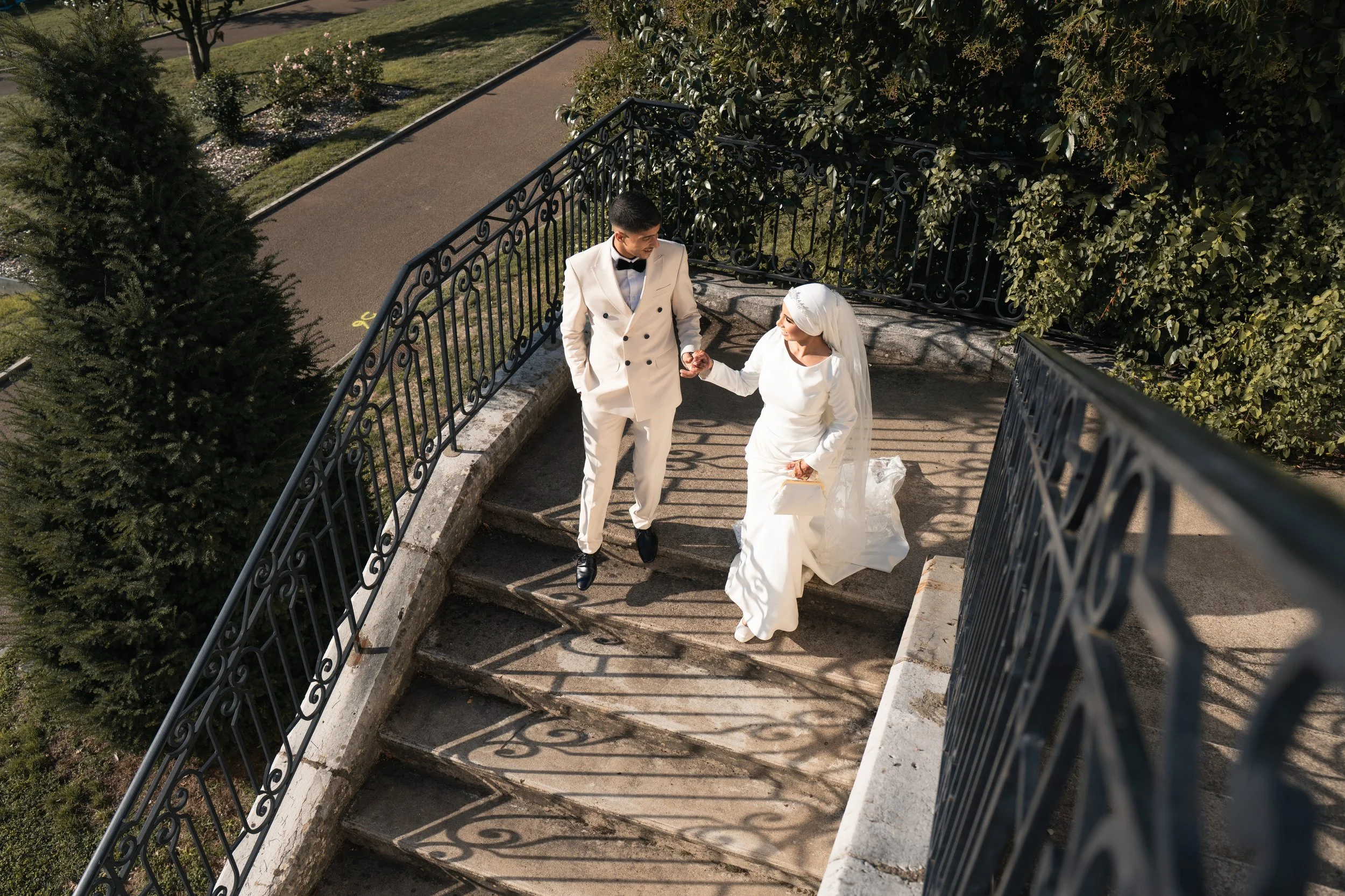 Un couple habillé en tenue de mariage descend des escaliers en pierre, avec des balustrades en fer forgé, dans un cadre extérieur verdoyant et ensoleillé.