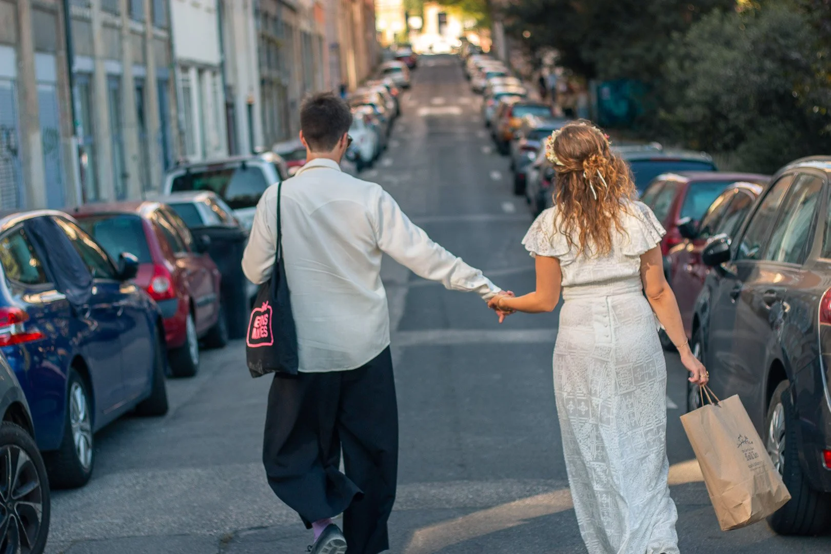Un couple se tient la main en marchant dans une rue urbaine bordée de voitures, on peut voir un homme et une femme tenant la main, la femme portant une robe blanche avec un sac de courses en papier.