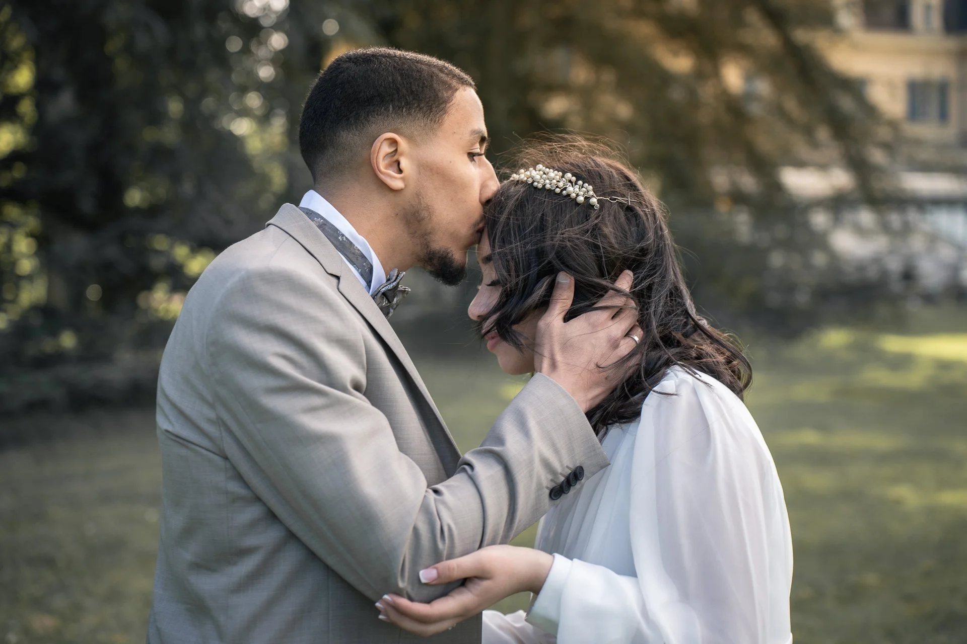 Un couple en tenue de mariage s'embrassant tendrement dans un parc, l'homme porte un costume gris clair et la femme une robe blanche avec une broche en perles dans les cheveux.