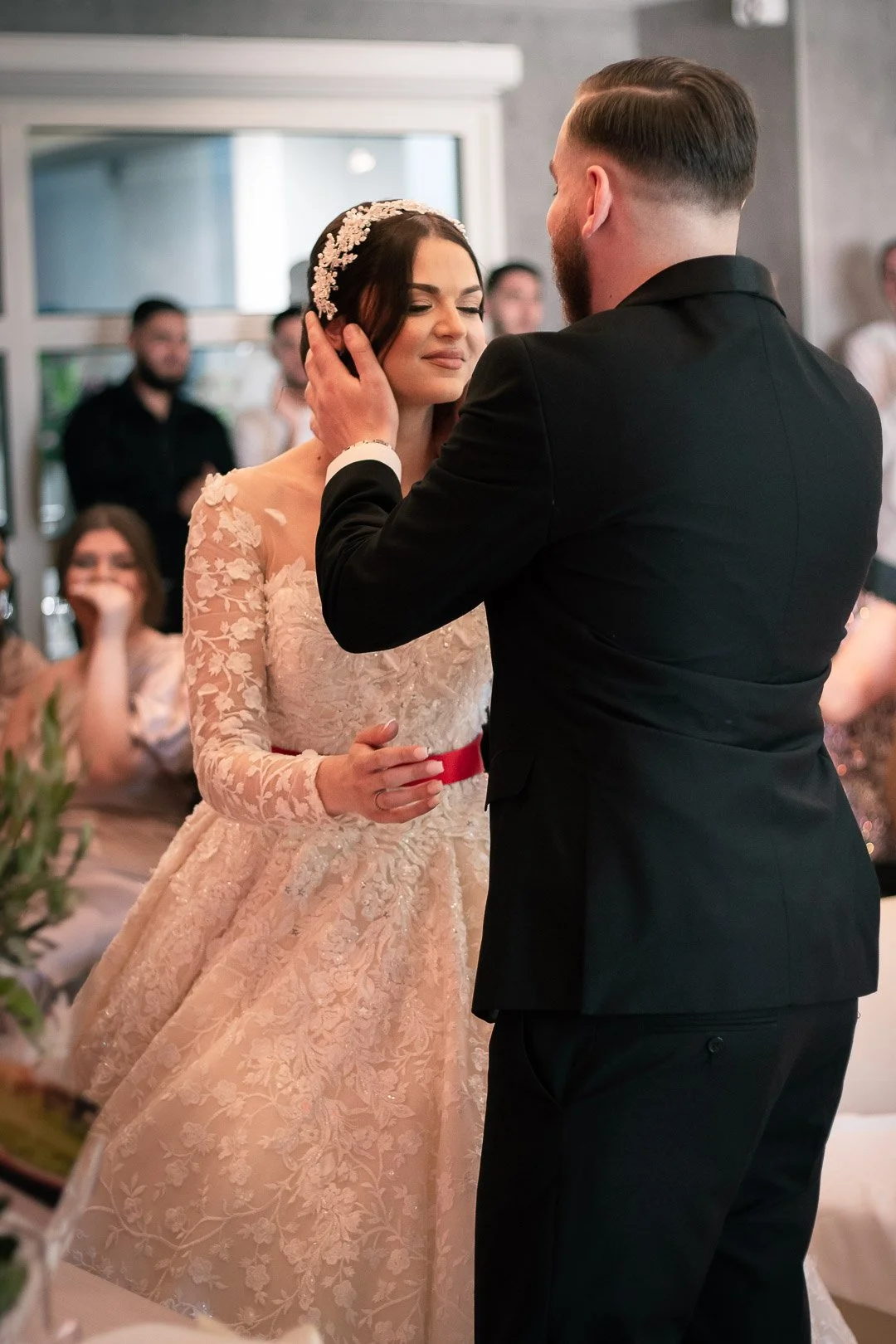Un couple lors de leur mariage, où l'homme semble échanger une promesse ou un rituel avec la femme, qui porte une robe de mariée élégante. Des invités sont visibles en arrière-plan, assis à une table.