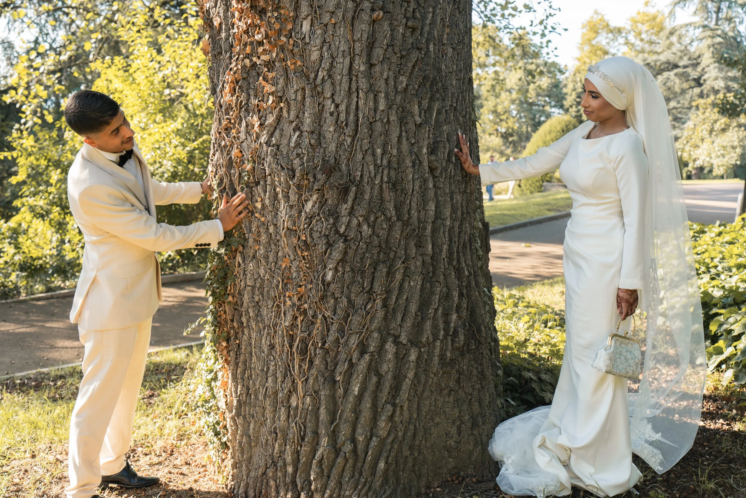 Un couple en tenue de mariage s'embrassant derrière un arbre dans un parc, avec des feuilles vertes et un ciel ensoleillé.