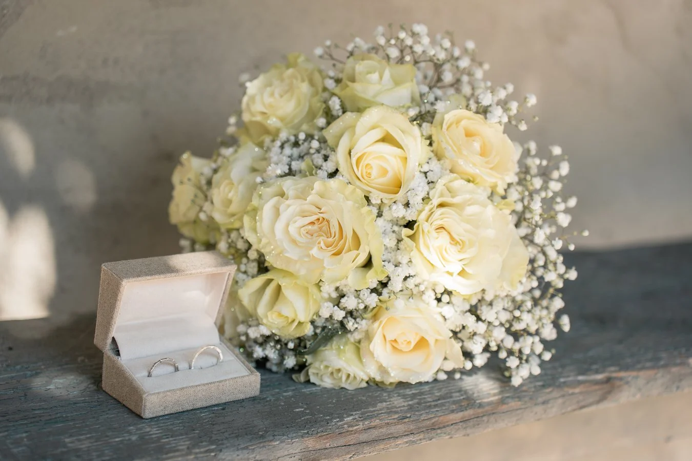 Un bouquet de roses blanches avec des petites fleurs blanches et deux alliances dans une boîte en velours gris, posé sur une table en bois.
