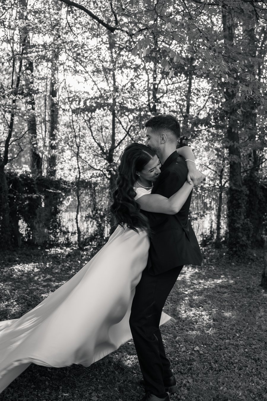 Un couple en mariage, en noir et blanc, dans une forêt, l'homme tenant la femme dans ses bras, tous deux souriant et riant.