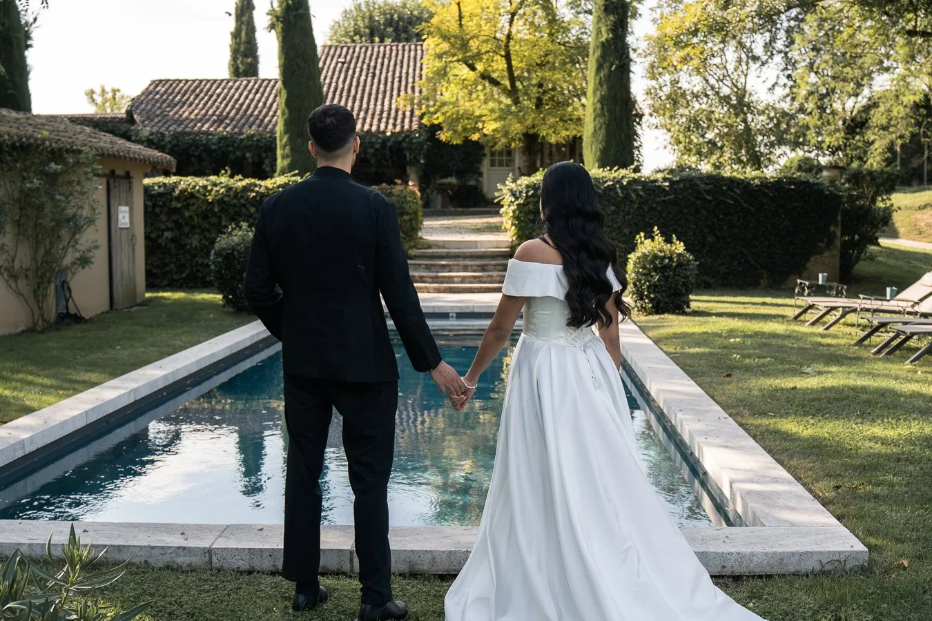 Un couple vêtu de habits de mariage, la femme en robe blanche et l'homme en costume noir, se tient par la main devant une piscine dans un jardin verdoyant, en arrière-plan une maison avec un toit en tuiles et de grands arbres en plein été.