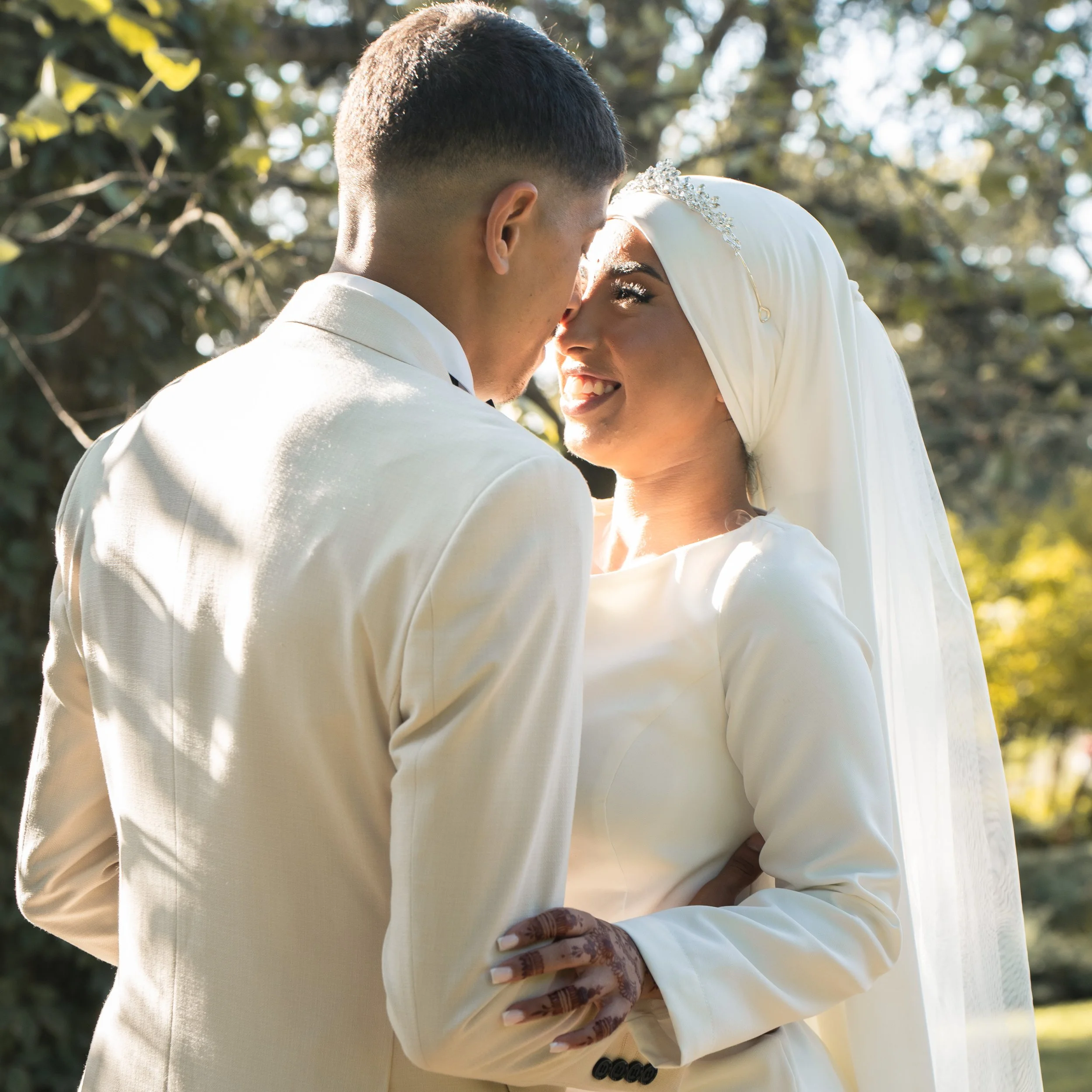 Un couple lors de leur mariage, un homme en costume blanc et une femme en hijab blanc souriant dans un environnement extérieur avec des arbres en arrière-plan.