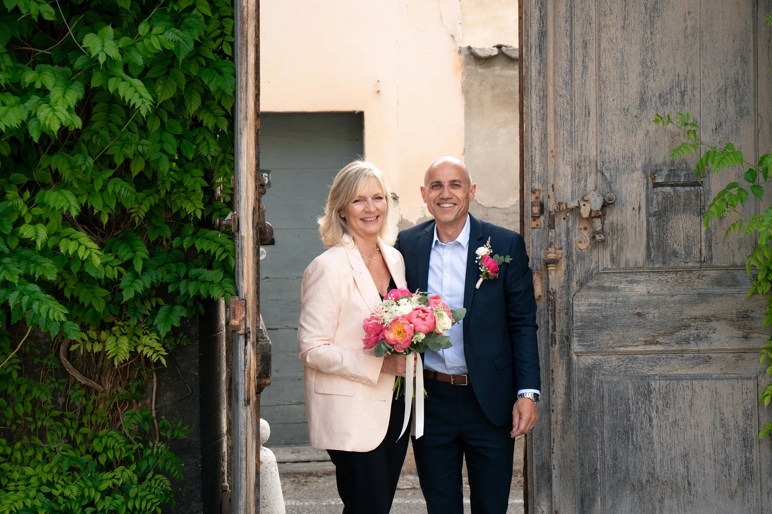 Un couple souriant, un homme et une femme, se tiennent à la porte d'une vieille bâtisse en bois, la femme tient un bouquet de fleurs, ils semblent heureux et élégants, entourés de végétation verte.