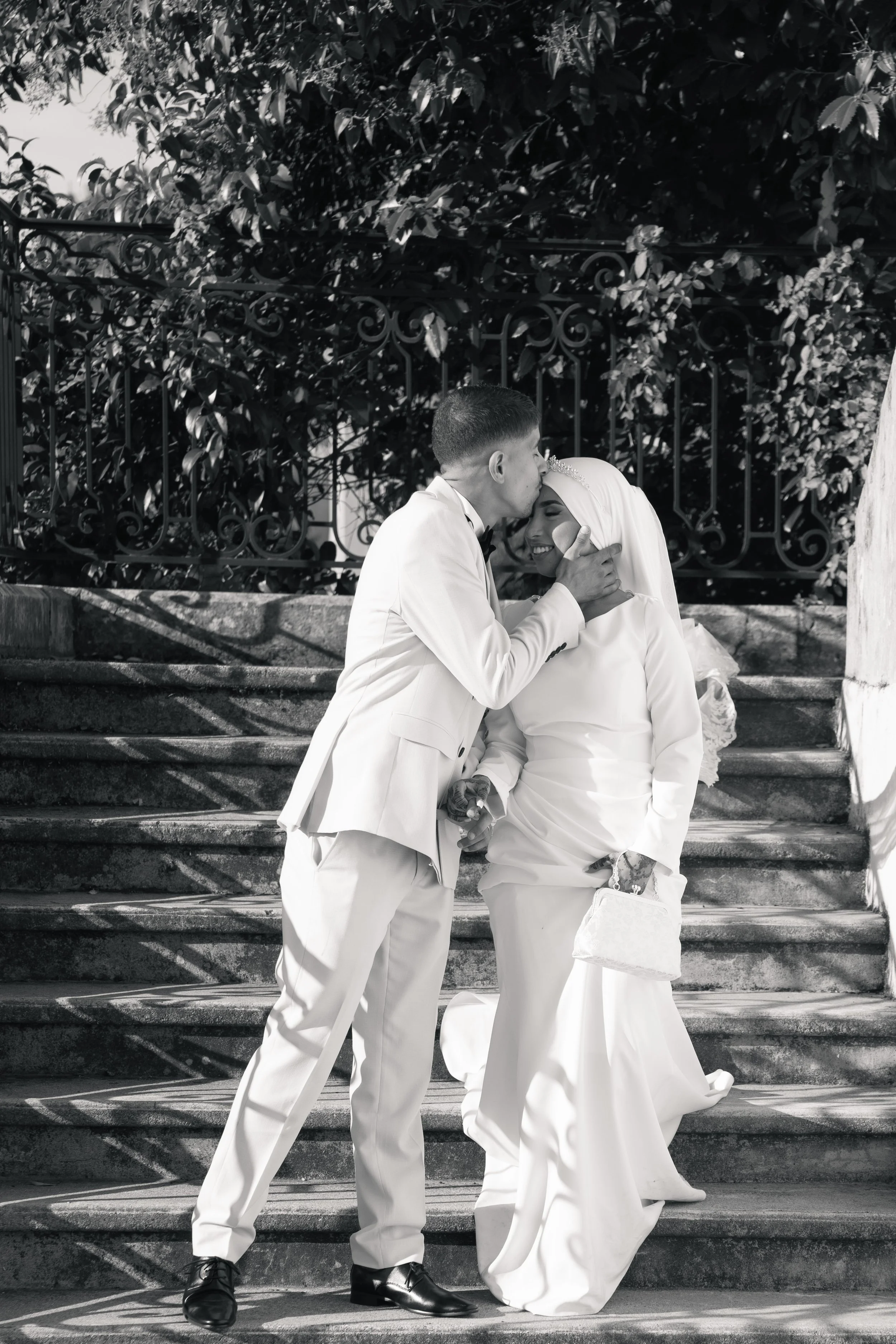 Un couple de mariés en costume blanc, la femme portant une robe de mariée, s'embrassent tendrement dans les escaliers en pierre, entourés d'un décor de feuillage et d'une barrière en fer forgé.