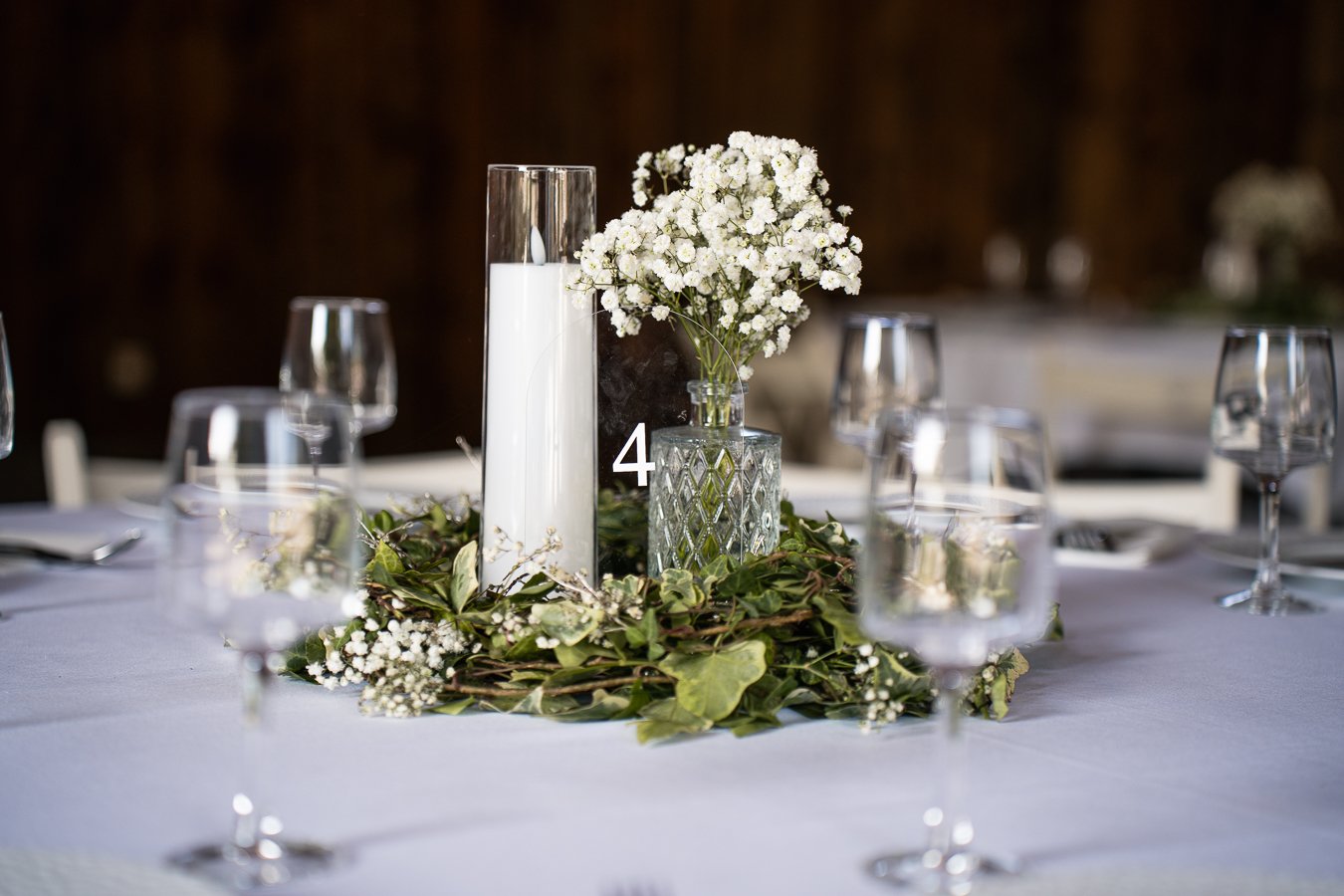 Table de mariage décorée avec un centre de fleurs blanches et des bougies dans un vase en verre, entourée de vaisselle en verre et de couverts, avec un fond obscur.
