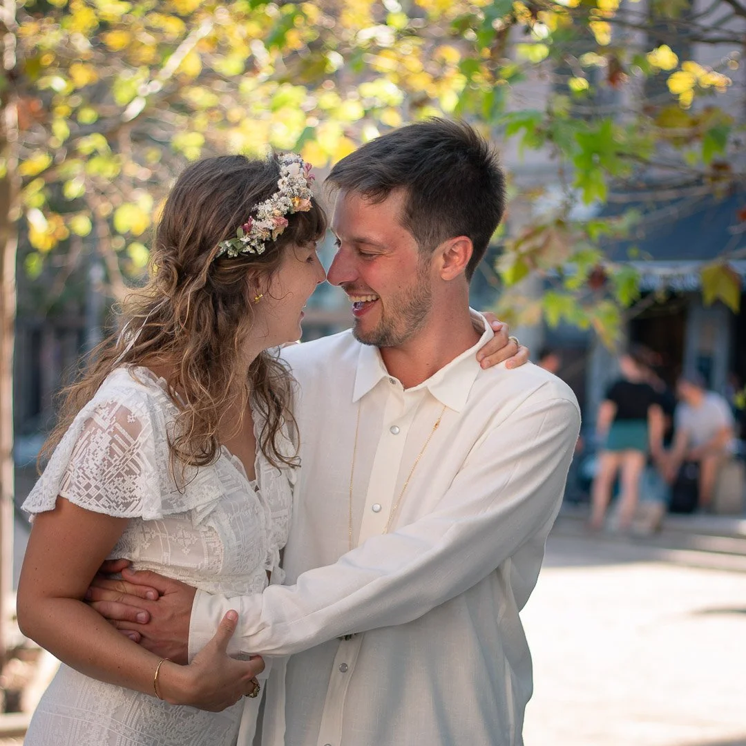 Un couple souriant en tenues blanches, peut-être lors d'une cérémonie ou d'une célébration, en plein air avec des arbres en arrière-plan.