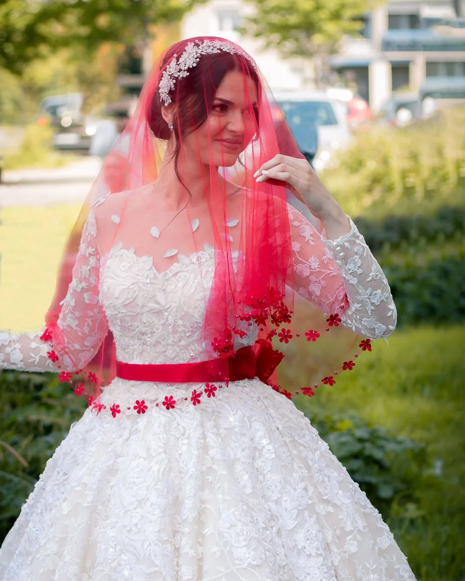 Une femme en robe de mariée blanche avec un voile rouge en tissu transparent et une décoration florale, tenant sa main près du visage, en extérieur avec des voitures en arrière-plan.