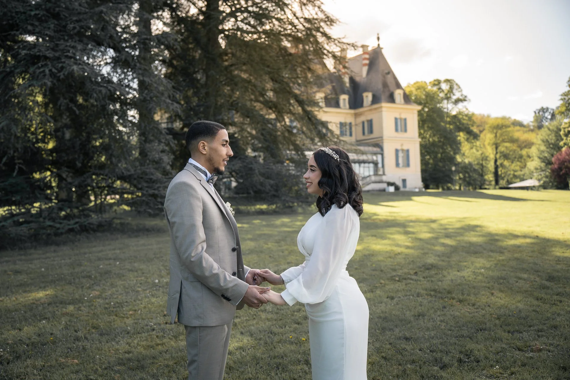 Un couple en vêtements de mariage se tenant la main dans un jardin devant un grand bâtiment avec une tour en arrière-plan, en pleine journée ensoleillée.
