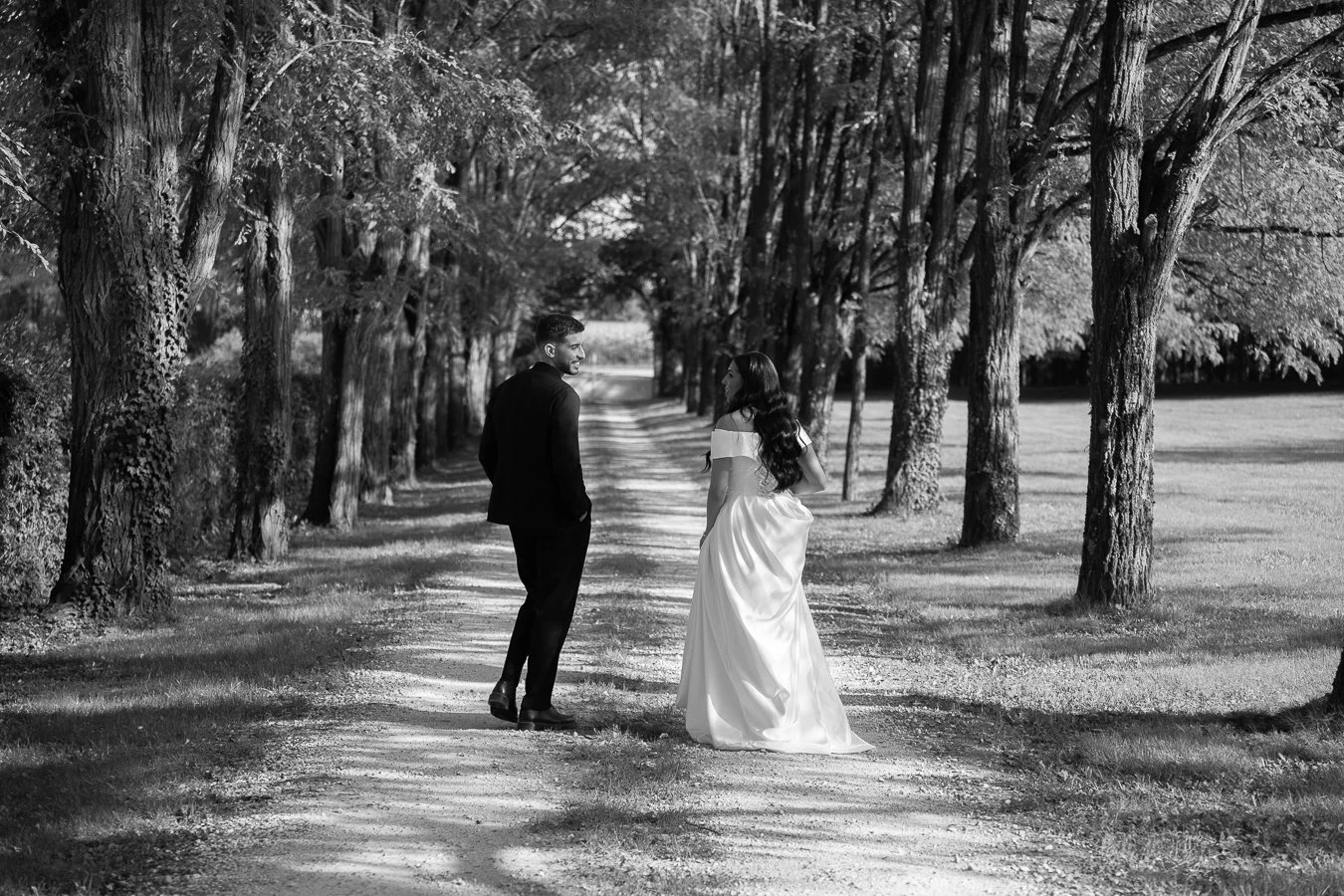 Un couple habillé en costume et robe de mariage marchant dans un chemin bordé d'arbres dans un parc, en noir et blanc.
