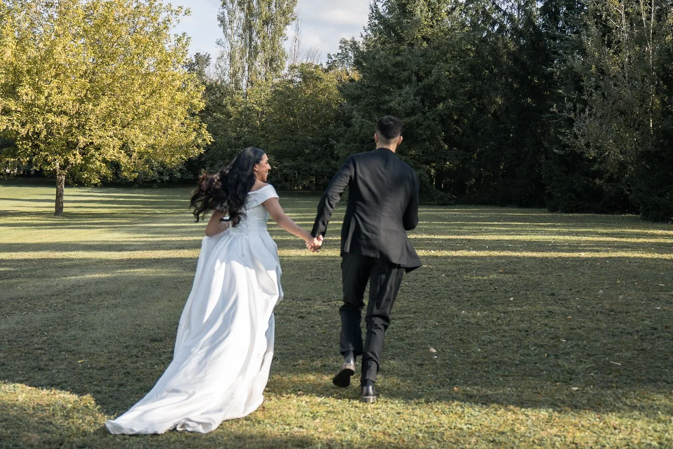 Un couple en costume de mariage se tient par la main dans un parc verdoyant, entouré d'arbres, lors d'une journée ensoleillée.