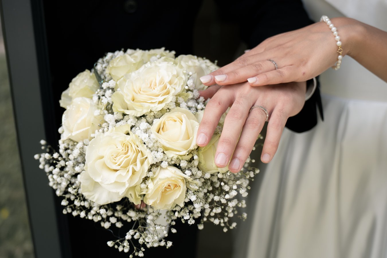 Mains de mariée montrant ses alliances, tenant un bouquet de roses blanches et de fleurs délicates.