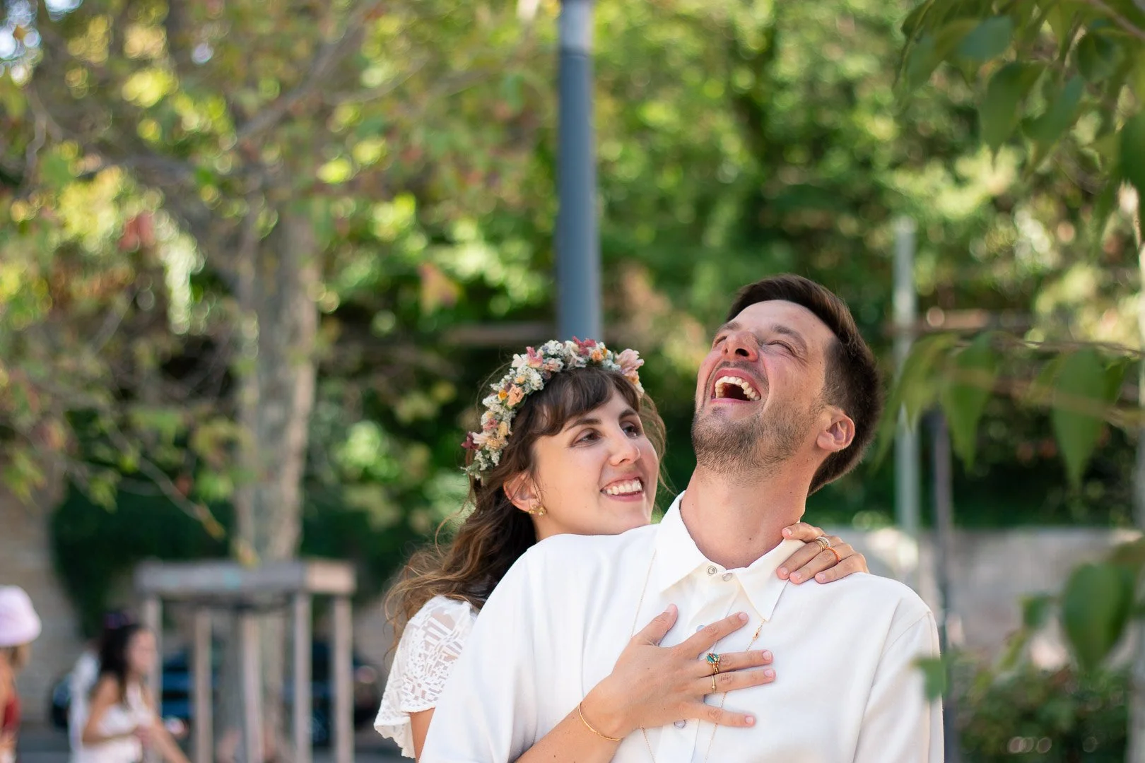 Un couple heureux, l'homme et la femme, en train de rire lors d'une célébration en plein air avec un décor naturel verdoyant. La femme porte une couronne de fleurs et l'homme un vêtement blanc.