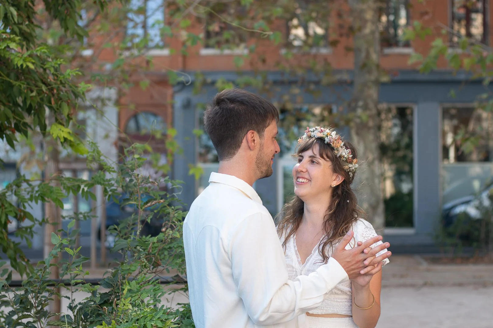 Un couple en vêtements blancs danse et regarde dans les yeux dans un cadre extérieur avec des arbres et un bâtiment en arrière-plan.