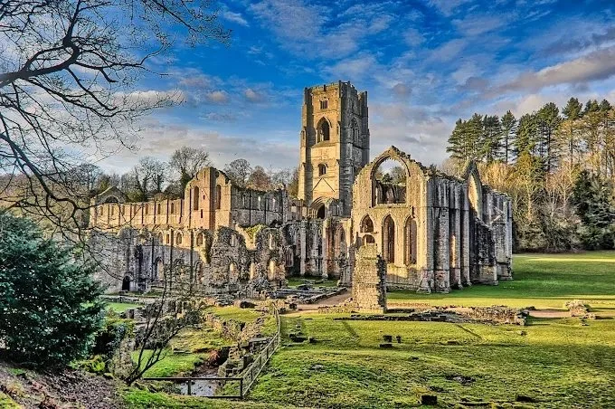 Ruins of an ancient stone building with a tower, surrounded by grassy area and trees, under a partly cloudy sky.