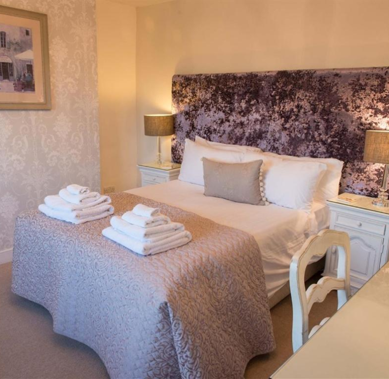 A neatly made hotel bed with white sheets, a gray decorative pillow, and two stacks of folded white towels on a textured bedspread, flanked by matching nightstands with lamps.
