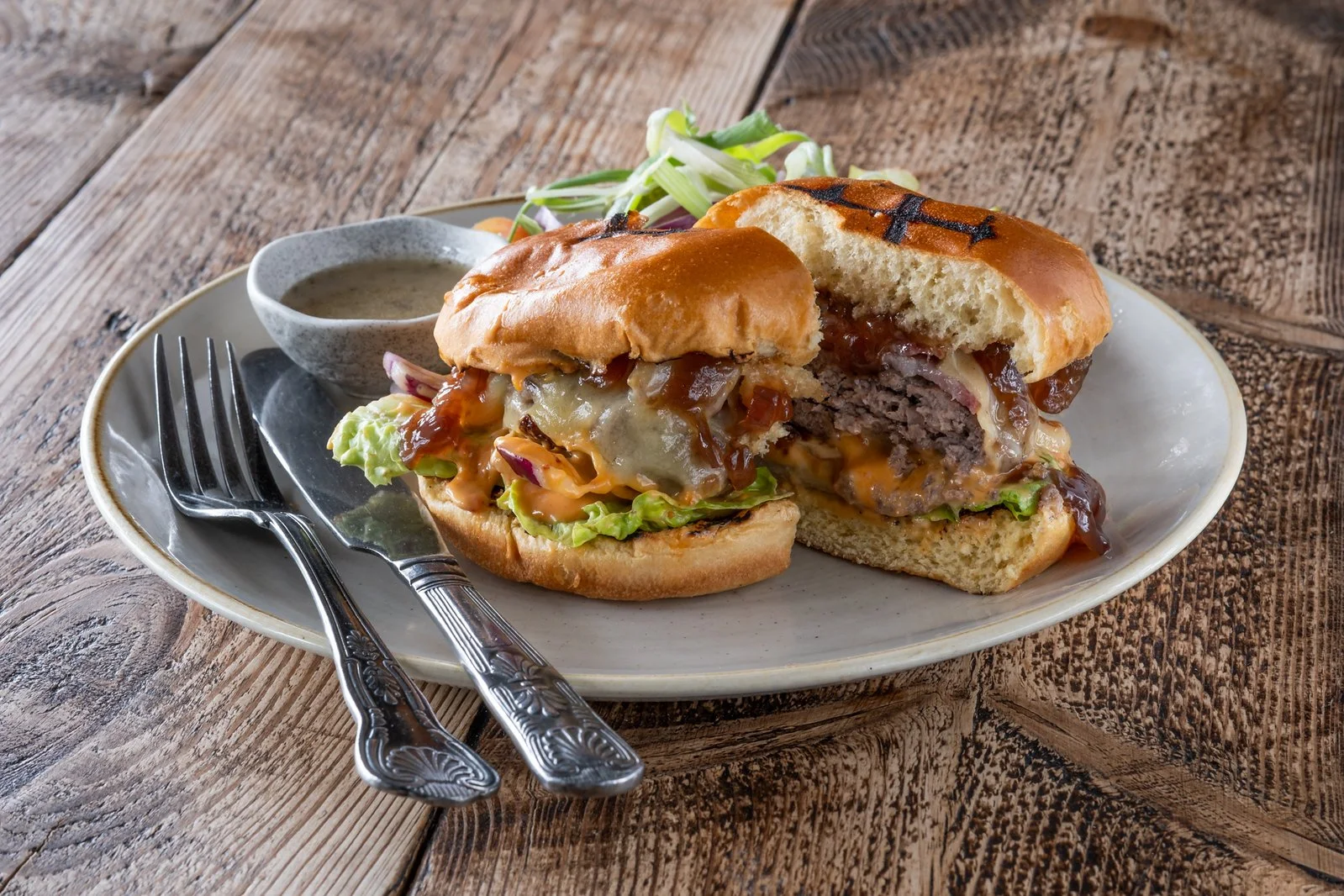 Half eaten cheeseburger with lettuce, onions, cheese, and beef patty on a white plate, with a side of condiments and salad, on a wooden table.