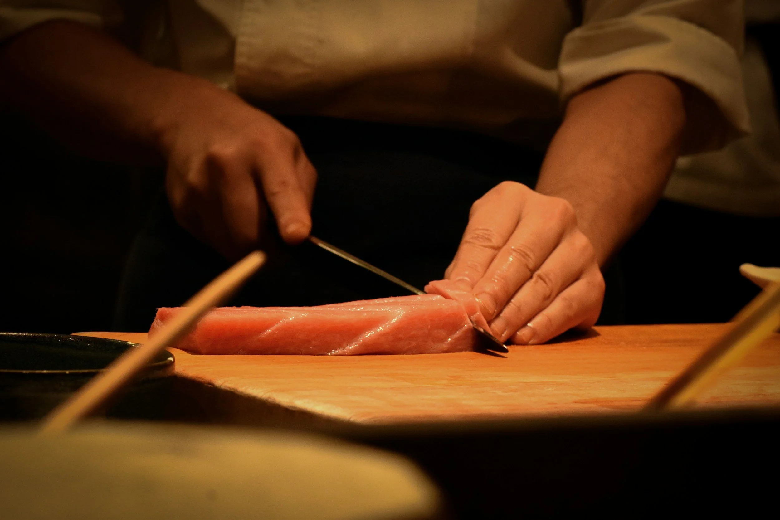 Person preparing raw fish, possibly sashimi, on a wooden cutting board in a dimly lit environment.